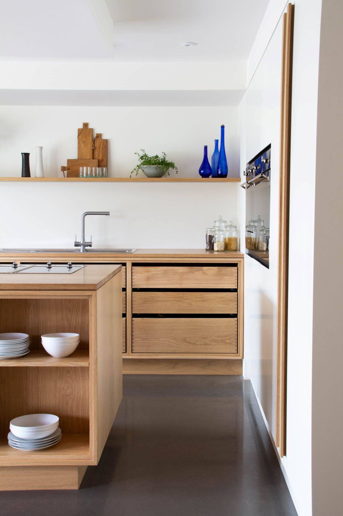 Minimalist kitchen with wooden cabinets and shelving. Open shelves display cutting boards, blue vases, and a small plant. The island has stacked bowls, and the countertop features jars. The space has a sleek, modern design with natural wood elements.