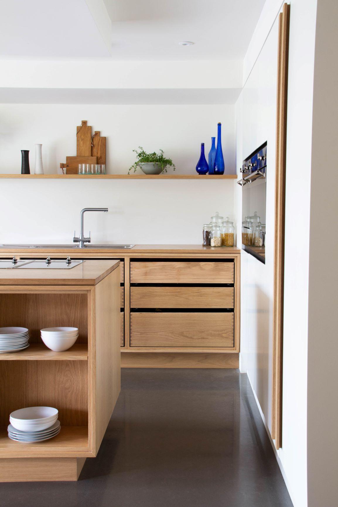 Minimalist kitchen with wooden cabinets and shelving. Open shelves display cutting boards, blue vases, and a small plant. The island has stacked bowls, and the countertop features jars. The space has a sleek, modern design with natural wood elements.