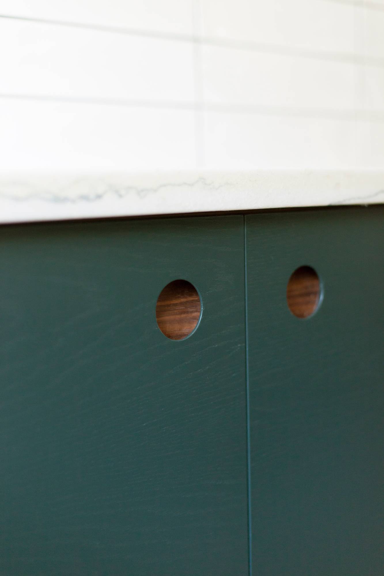 Close-up of dark green cabinet doors with round cut-out handles, topped with a white marble countertop. The background features white tiled walls.