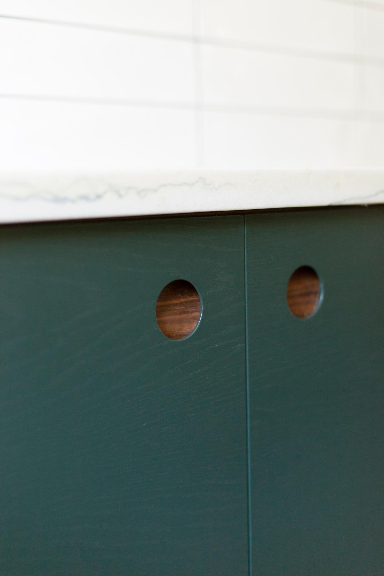 Close-up of dark green cabinet doors with round cut-out handles, topped with a white marble countertop. The background features white tiled walls.