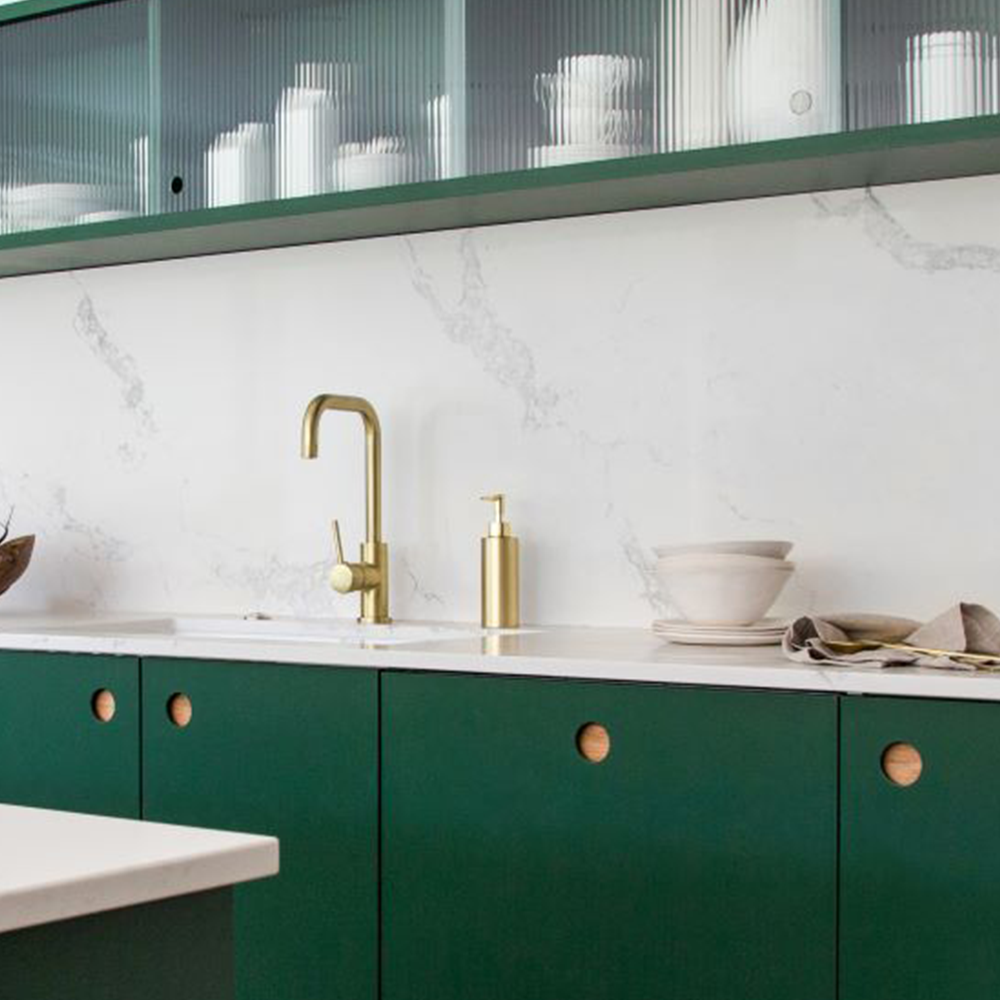 A modern kitchen with green cabinets, a gold faucet, white marble backsplash, and white countertops. Upper cabinets have glass doors displaying white dishes, and there are minimalistic accessories on the counter.