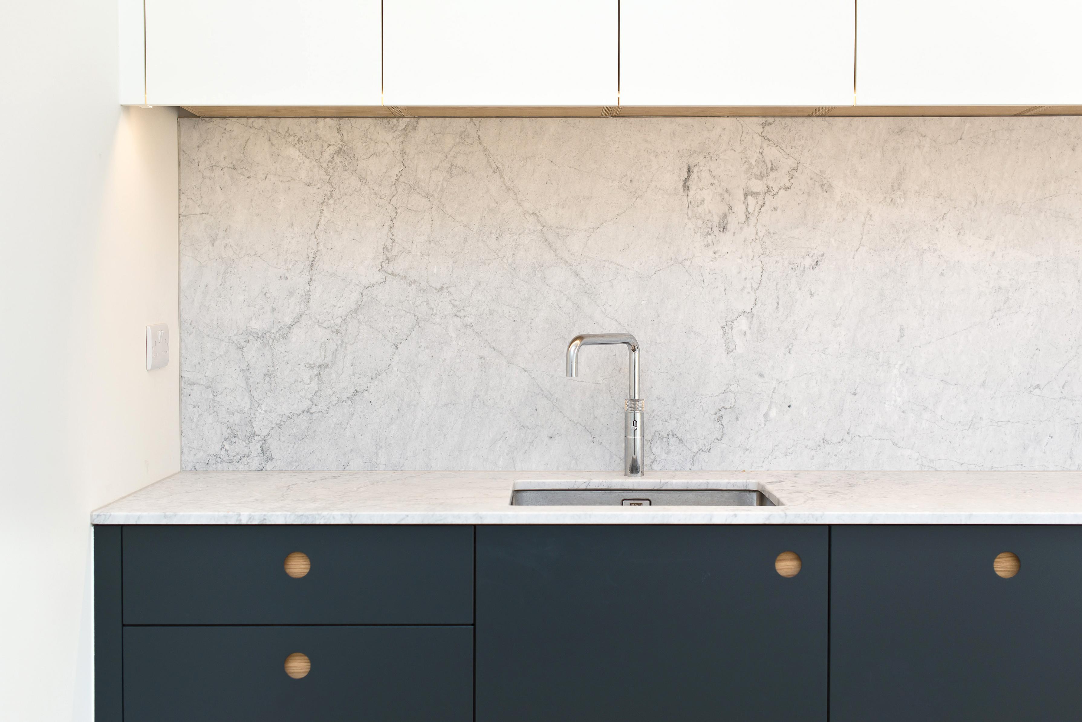 A modern kitchen counter featuring dark teal cabinetry with circular wood-inlaid cutout handles, a white marble countertop and matching high splashback, a stainless steel sink and tap, and minimalist white upper cabinets.