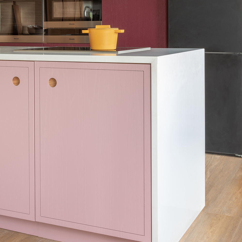 A modern kitchen with pink cabinets, a white countertop, a yellow pot on top, wooden flooring, and built-in ovens reflected in a glass surface.