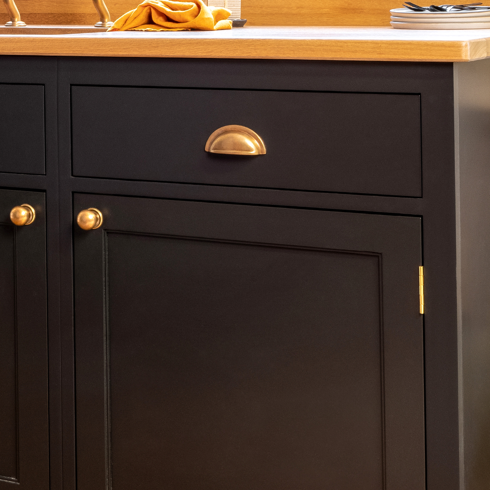 Close-up of a black kitchen cabinet with gold handles and a wooden countertop. A stack of plates and a folded yellow cloth are on the counter in the background.