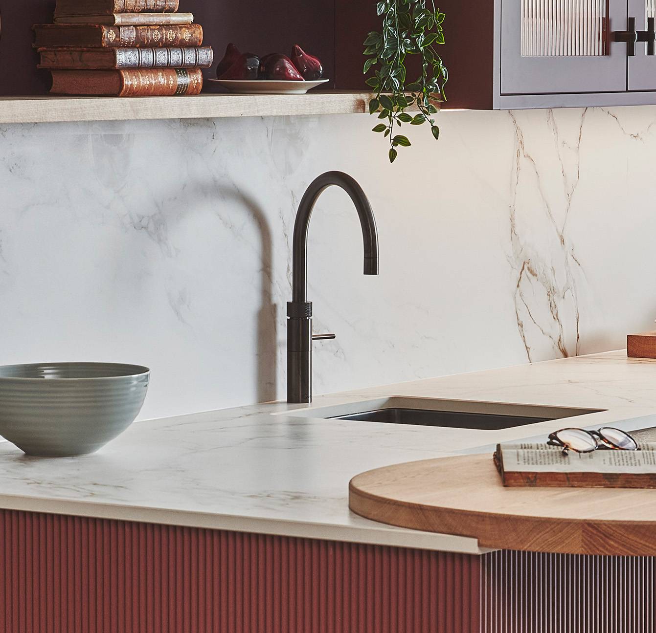 Modern kitchen with a marble countertop and backsplash. Features a sleek black faucet and an inset sink. Greenery hangs from above, and a stack of books rests on the shelf. A bowl and open magazine sit on the counter.