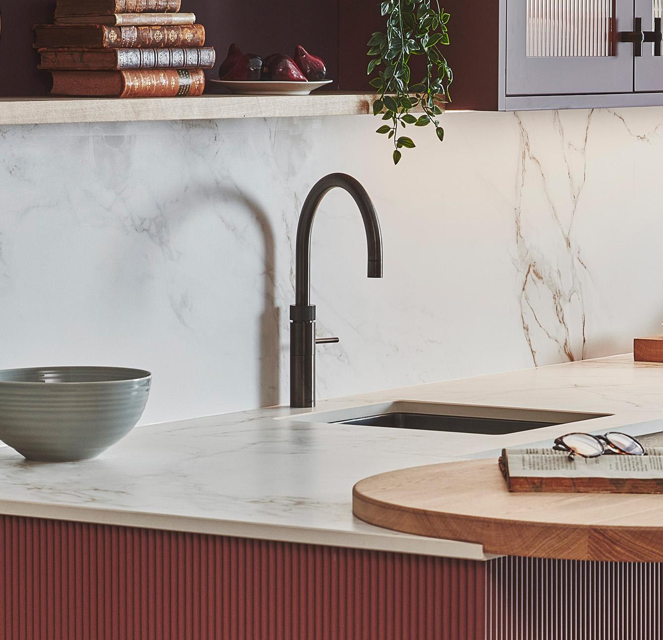 Modern kitchen with a marble countertop and backsplash. Features a sleek black faucet and an inset sink. Greenery hangs from above, and a stack of books rests on the shelf. A bowl and open magazine sit on the counter.