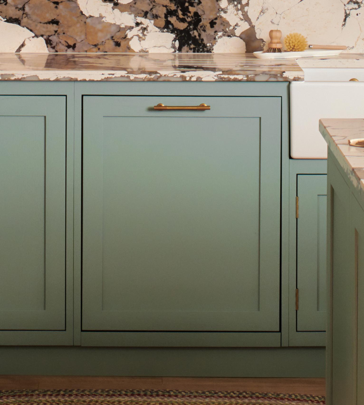 Close-up of a kitchen featuring sage green cabinets with brass handles, a white farmhouse sink, and a marble countertop. The backsplash displays a distinct, organic marble pattern in various shades of gray and beige.