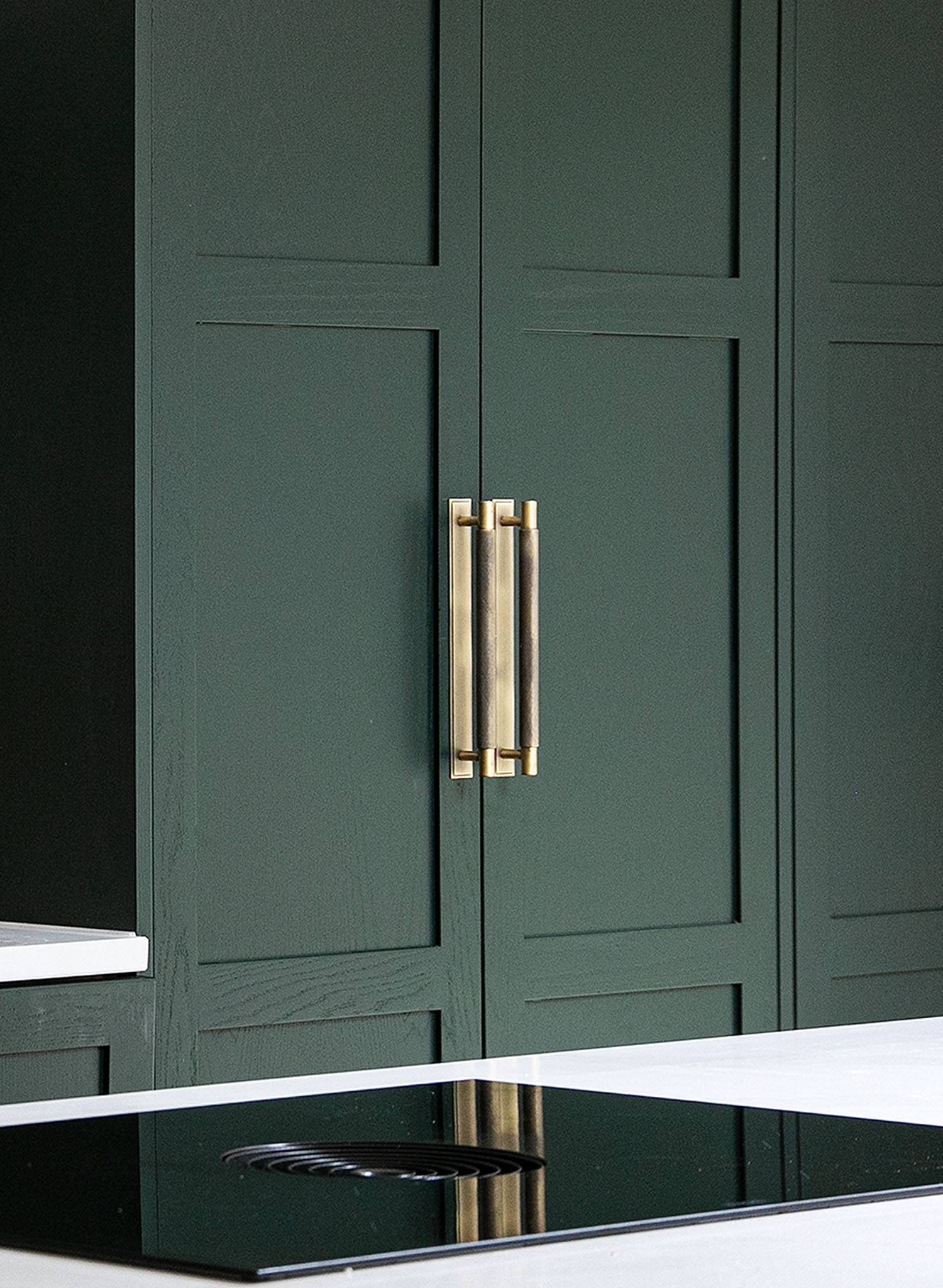 Dark green modern kitchen cabinets with brass handles, next to a white countertop and a black glass electric cooktop in the foreground.