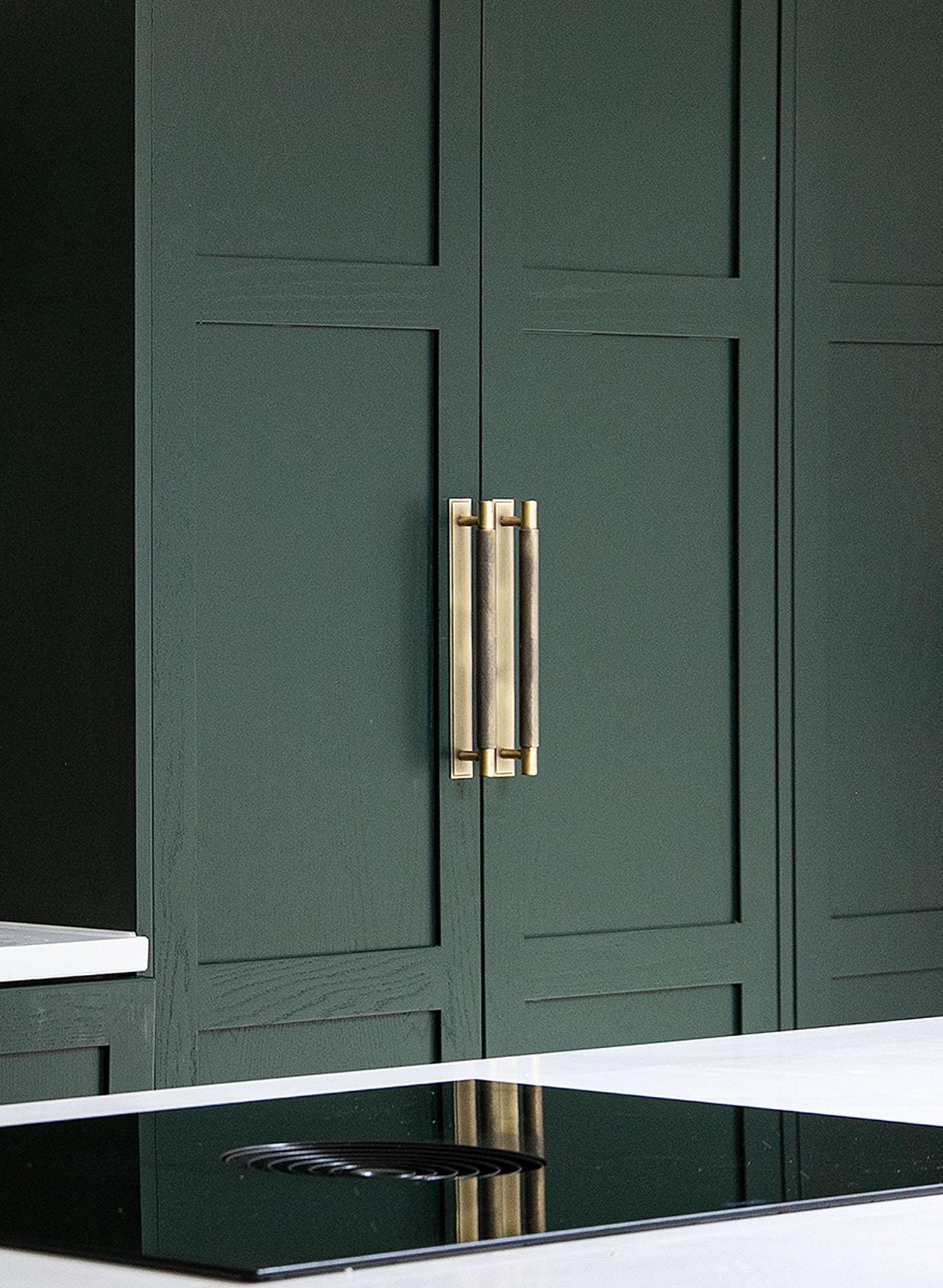 Dark green modern kitchen cabinets with brass handles, next to a white countertop and a black glass electric cooktop in the foreground.