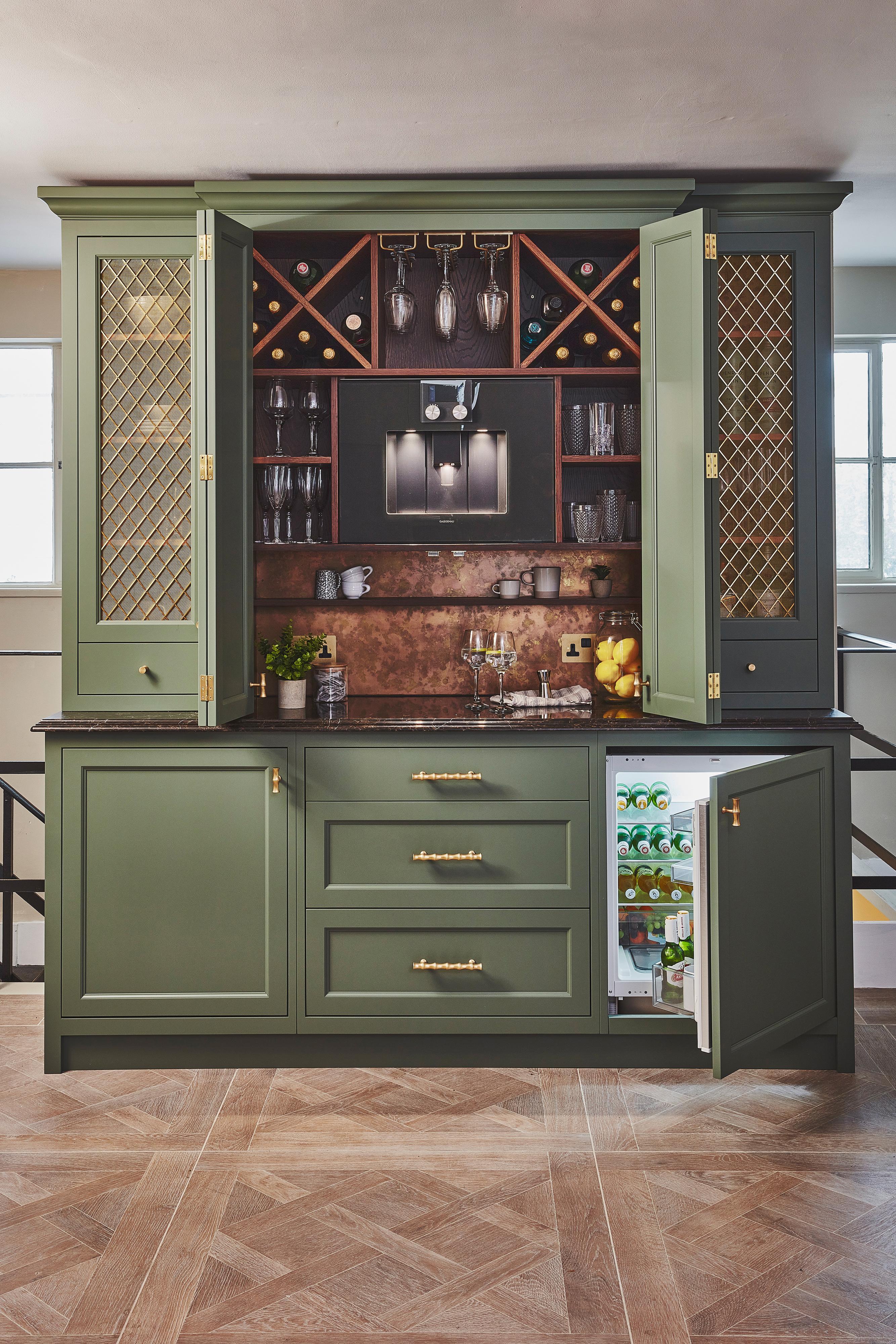 A stylish home bar with green cabinets, gold handles, a wine rack, glassware, and a mini fridge stocked with drinks. The countertop holds bottles, glasses, and bar tools. The cabinet doors are partly open.