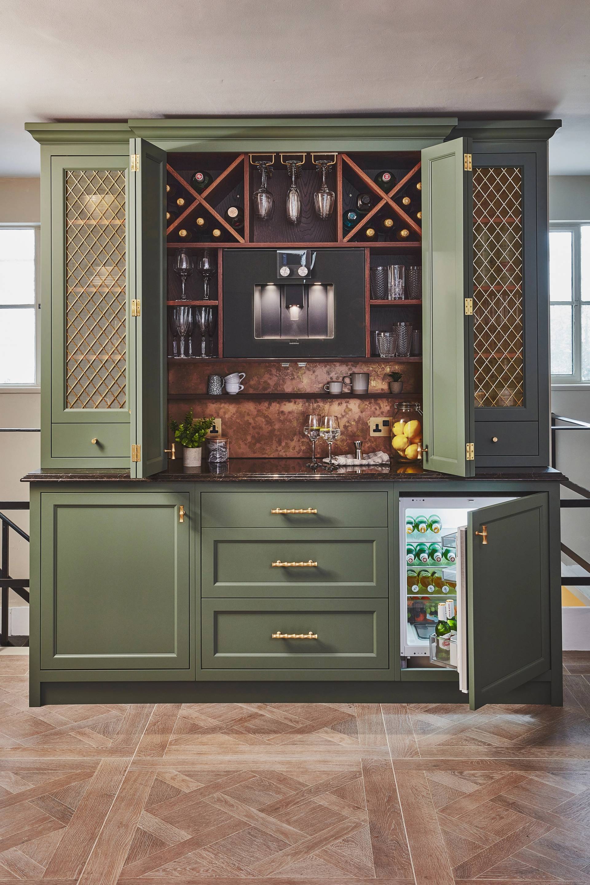 A stylish home bar with green cabinets, gold handles, a wine rack, glassware, and a mini fridge stocked with drinks. The countertop holds bottles, glasses, and bar tools. The cabinet doors are partly open.
