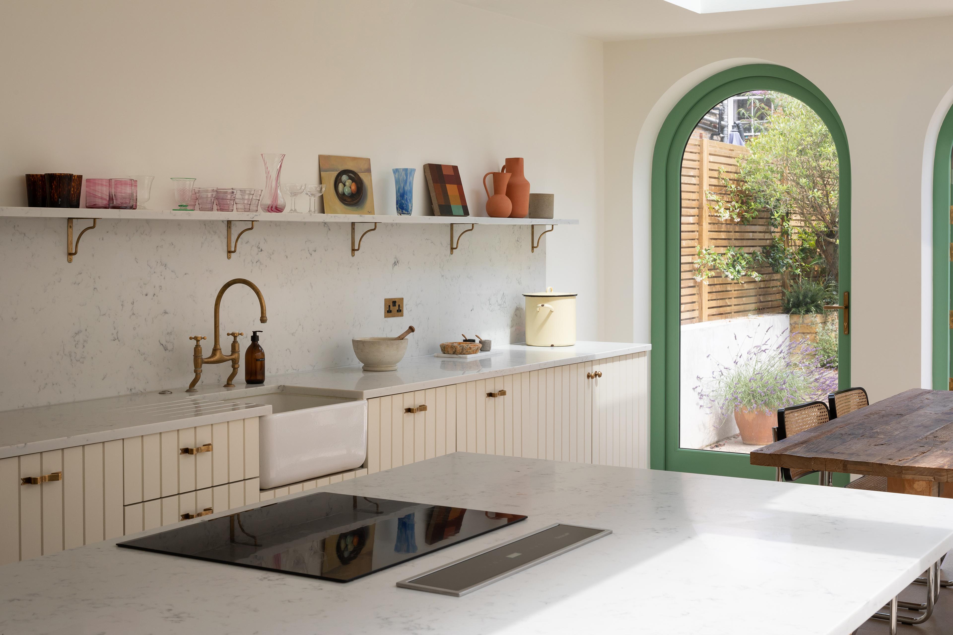 A bright kitchen with a marble countertop and brass faucet. Open shelves display dishes and decor. Large arched window and door with green frames lead to a garden. A wooden dining table is visible on the right. Light fills the space.
