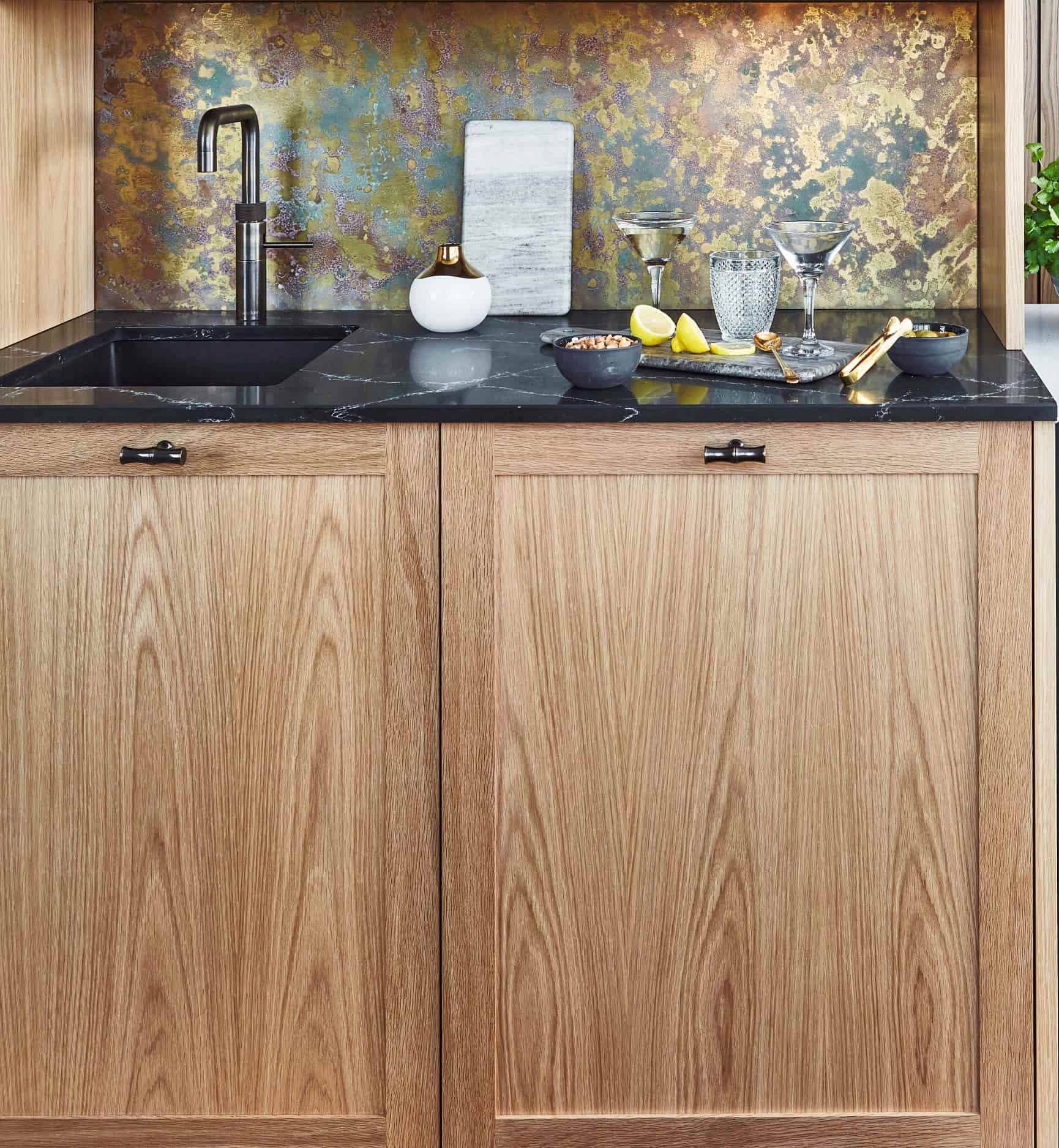 A modern kitchen sink with a black faucet, black countertop, light wood cabinets, and a metallic, speckled backsplash. On the counter are glasses, lemons, a bowl, utensils, and a decorative vase.