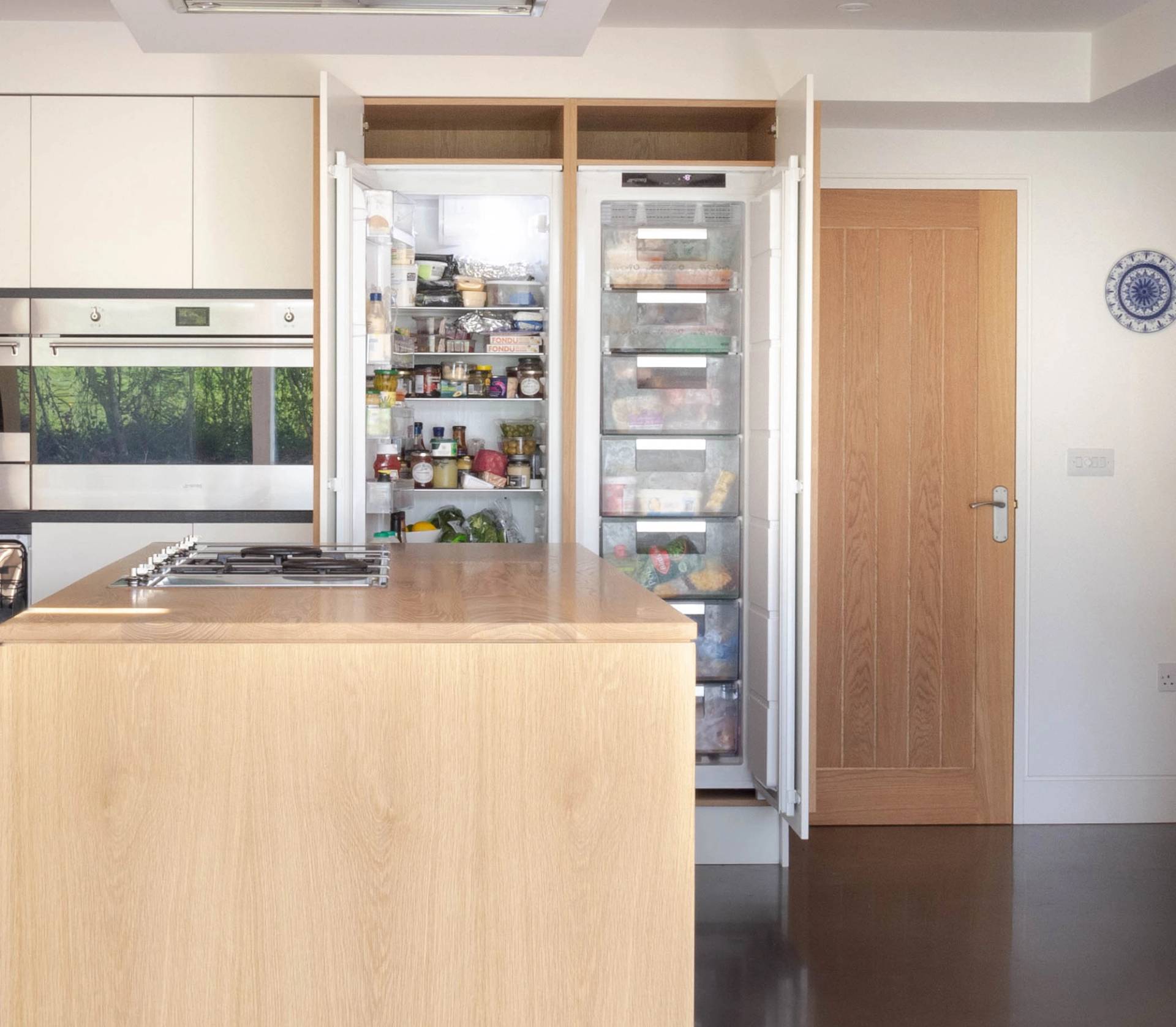 Modern kitchen with a large open refrigerator and freezer filled with various food items. Light wooden cabinets, a central island with a stove, and a closed wooden door are visible. A decorative plate is hung on the wall.