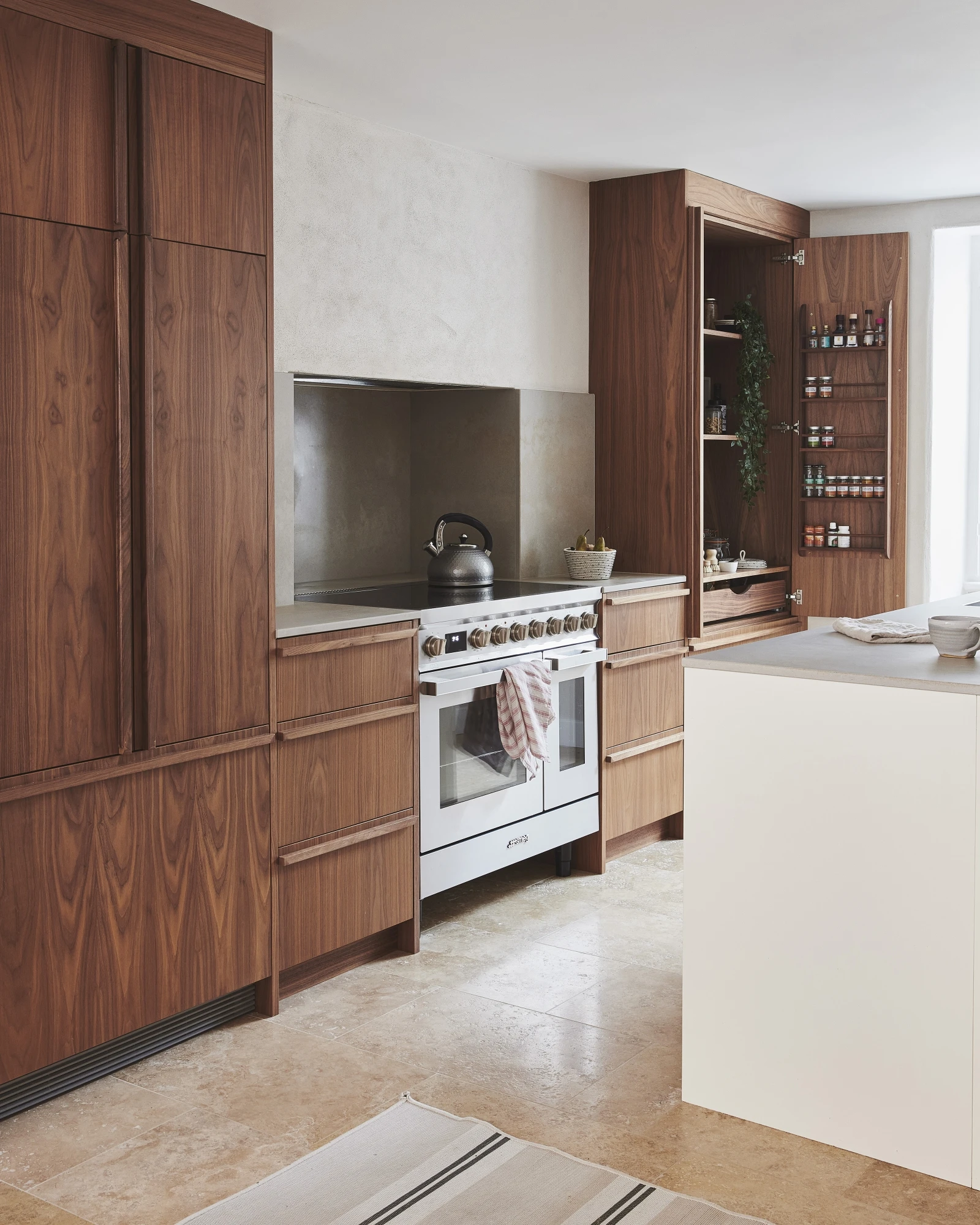 Modern kitchen with wooden cabinets featuring a white oven and stovetop. A kettle sits on the stove. Open shelving with spices and a plant is on the right. A central island with a light countertop is in the foreground, and a striped rug is on the floor.