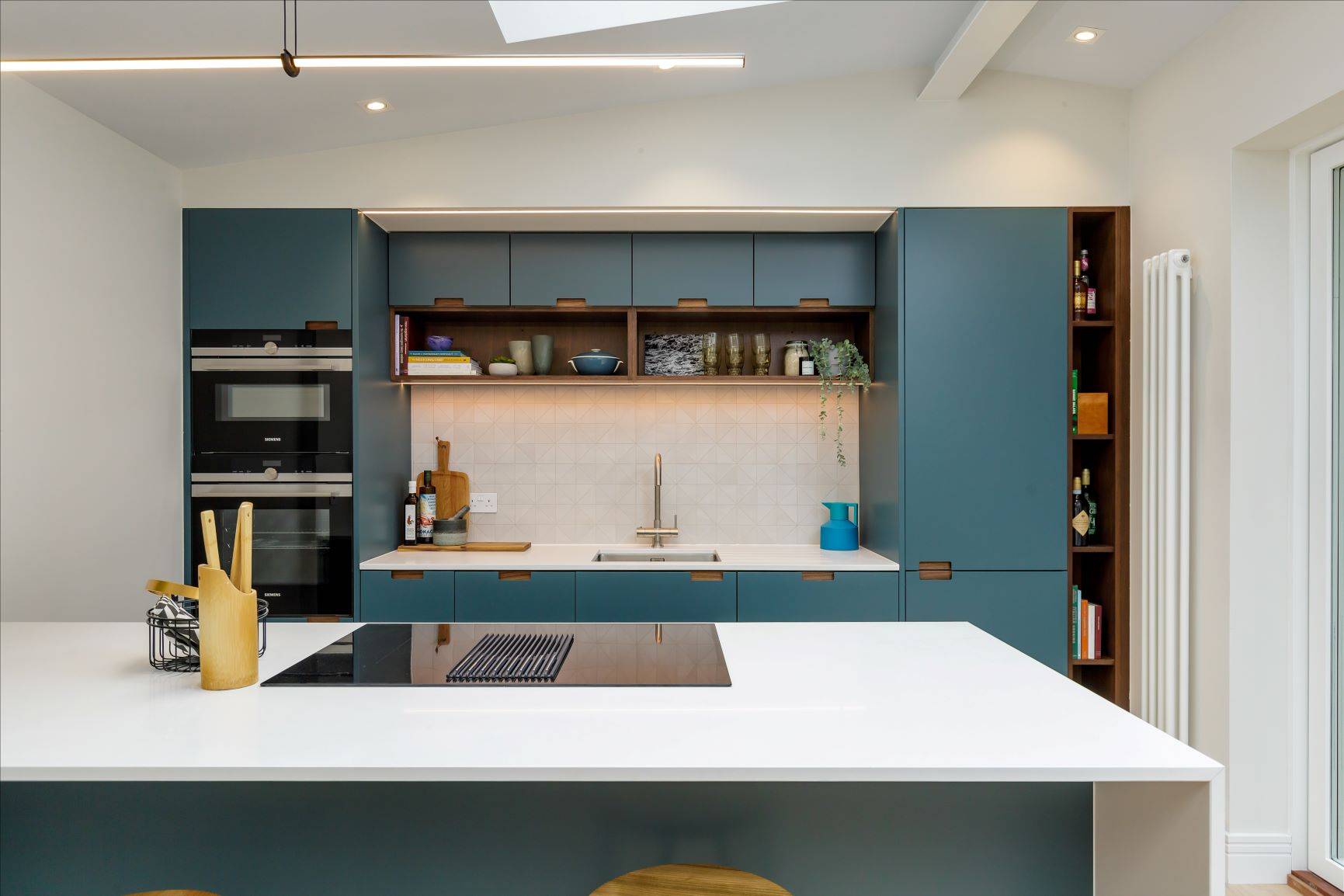 A modern kitchen featuring teal cabinets, a white countertop island with an induction cooktop, wooden stools, built-in appliances, and open shelving with décor and kitchenware. Soft lighting highlights the sleek design and clean lines.