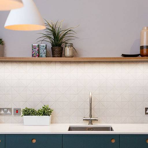Modern kitchen with a geometric-patterned white backsplash, a stainless steel sink, and a wooden floating shelf holding plants, jars, and mugs. A white pendant light hangs from the ceiling.