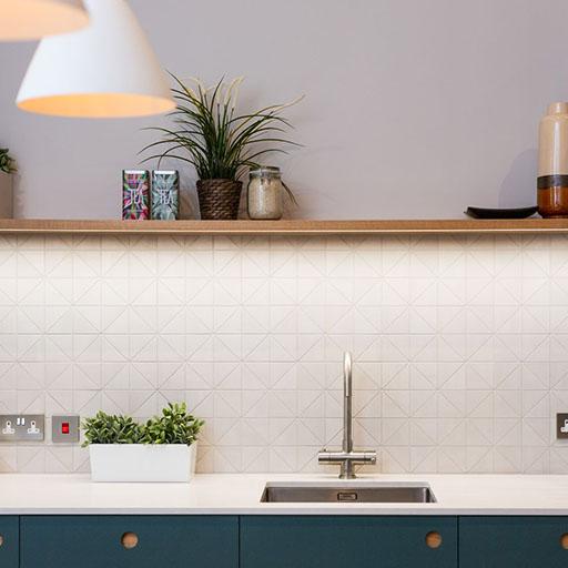 Modern kitchen with a geometric-patterned white backsplash, a stainless steel sink, and a wooden floating shelf holding plants, jars, and mugs. A white pendant light hangs from the ceiling.