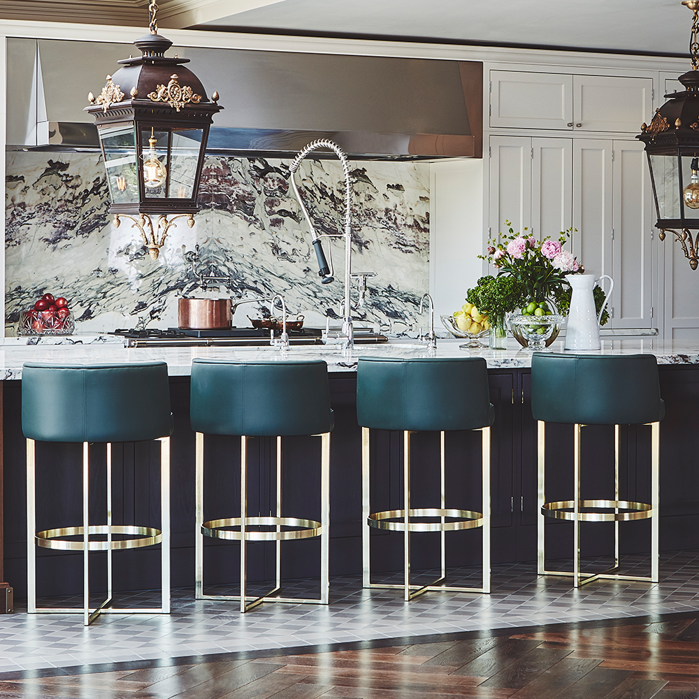 A modern kitchen with a marble island, four green and gold barstools, a marble backsplash, pendant lights, and fresh flowers on the counter. The cabinetry is white and the flooring is wood.