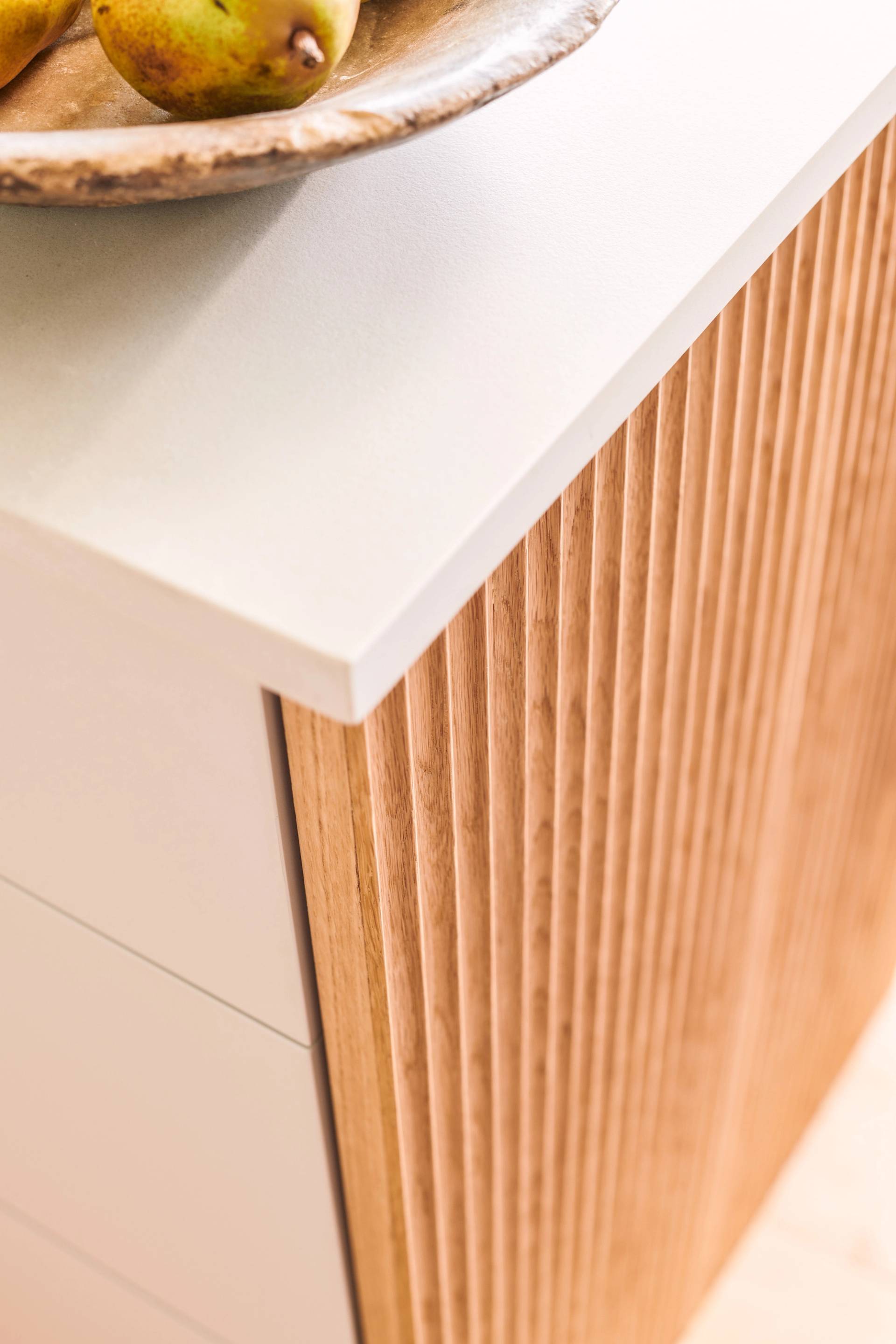 Close-up of a modern kitchen counter featuring a smooth white countertop and a vertical slatted wood cabinet. A wooden bowl with pears is partially visible on top of the counter.
