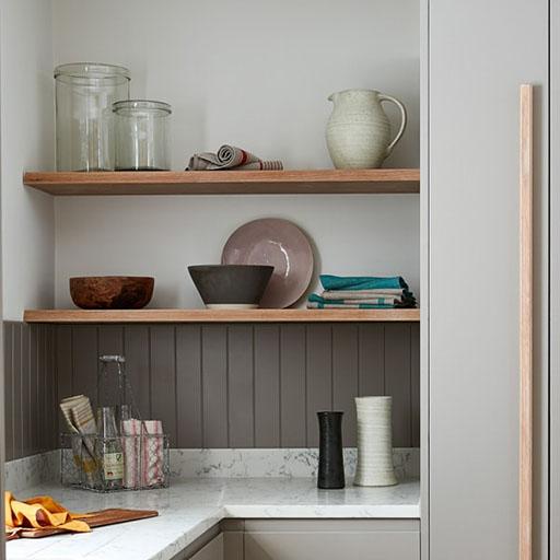 Minimalist kitchen shelves with glass jars, a ceramic jug, bowls, plates, and folded linens. Below, a marble countertop holds dish soap, a striped towel, and vases against a gray paneled backsplash.