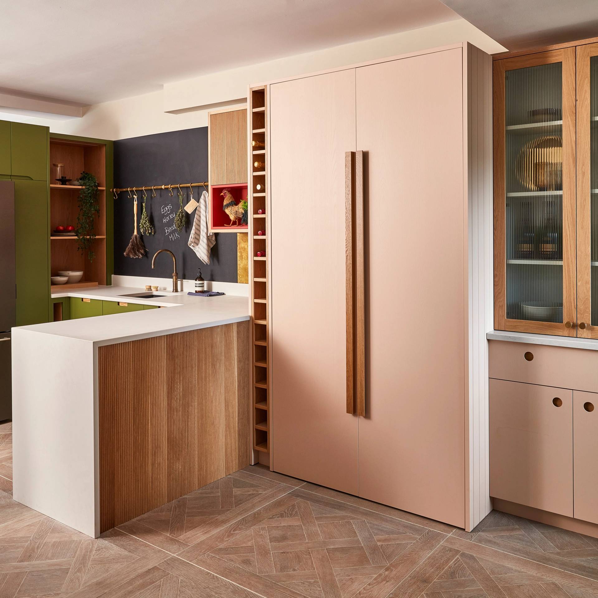 Modern kitchen with a pastel pink fridge, wooden cabinets, and green accents. A small island with a white countertop includes a sink and hanging plants. Light wood flooring and a black backsplash add contrast to the space.