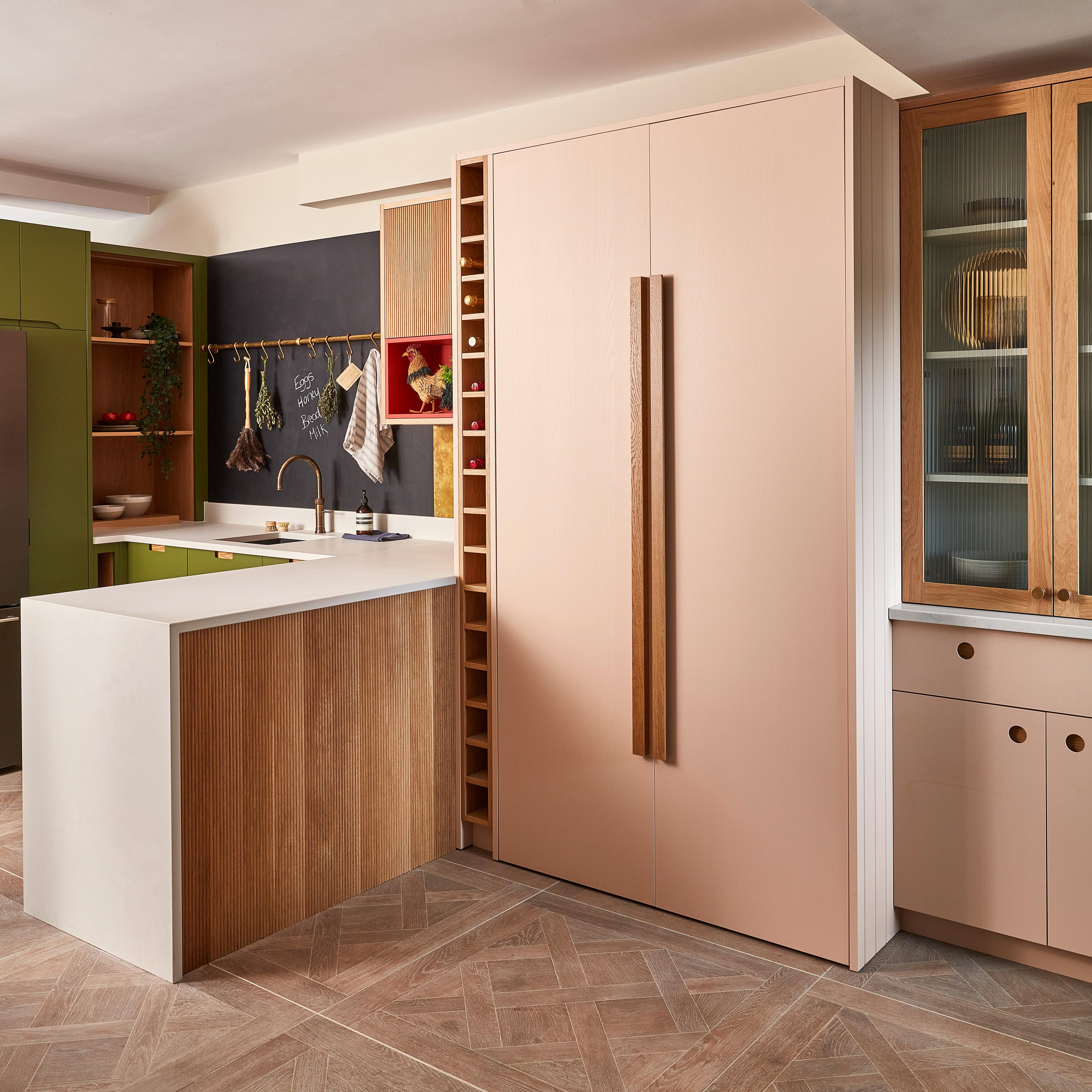 A modern kitchen with green and wood cabinets features a hidden home office nook with shelves, a computer, and a chair inside a cabinet that can be closed when not in use. There are neutral tones and potted plants for decor.