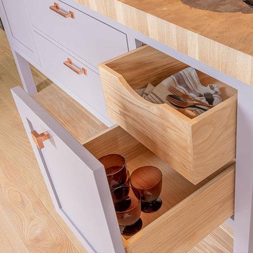 A wooden kitchen cabinet with two open drawers: the top drawer contains napkins and cutlery, while the bottom drawer holds several brown glasses. The countertop and floor are also wood.