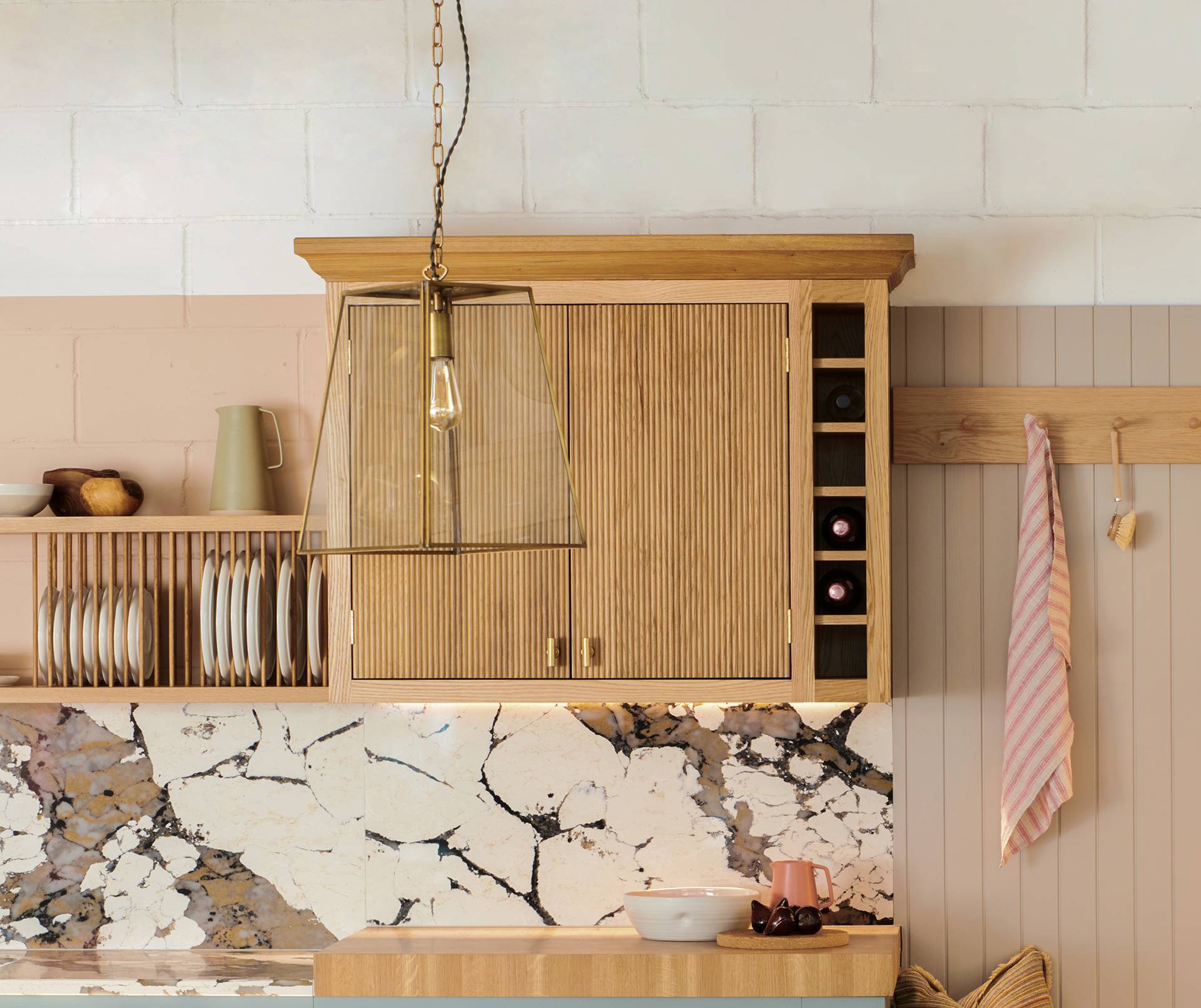 A cozy kitchen corner featuring light wooden cabinets, a marble backsplash, and a set of hanging cups. A modern pendant light hangs above a small countertop with kitchen items, and a striped towel is hung on the wall.