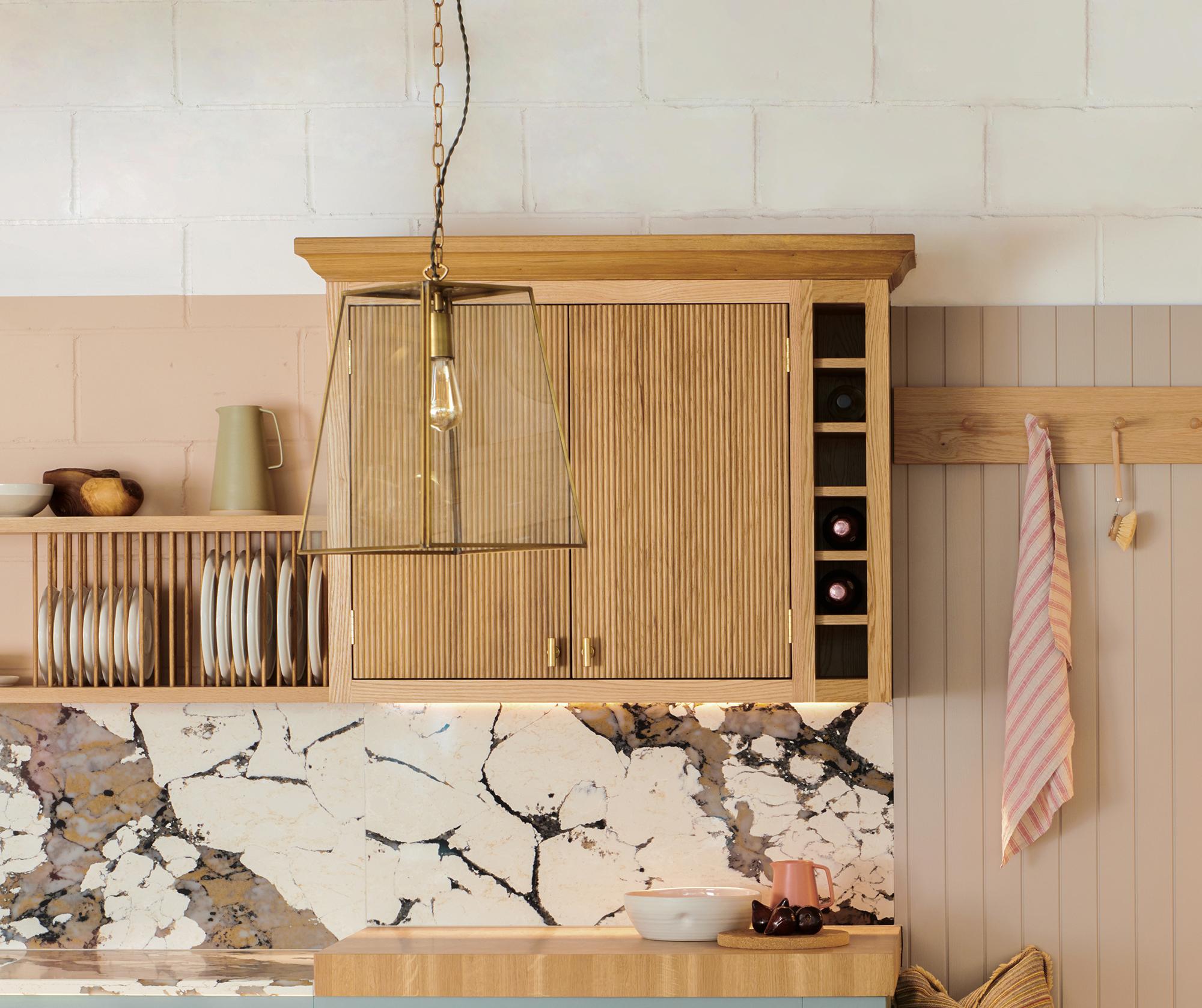 A cozy kitchen corner featuring light wooden cabinets, a marble backsplash, and a set of hanging cups. A modern pendant light hangs above a small countertop with kitchen items, and a striped towel is hung on the wall.