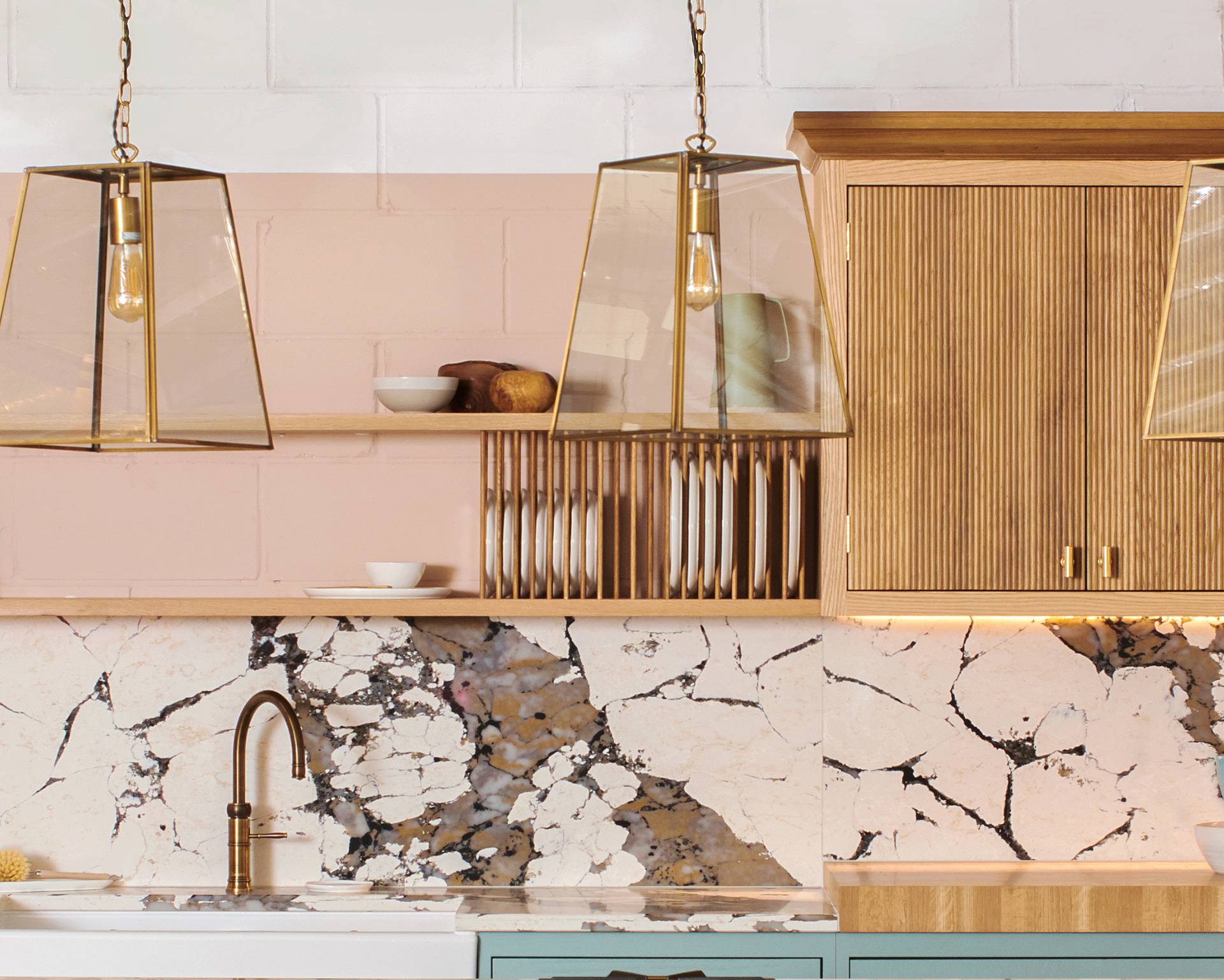 Modern kitchen with gold pendant lights, a marble-patterned backsplash, open wooden shelves holding dishes, a gold faucet, and light wood cabinets. The countertop is white with a light blue lower cabinet.