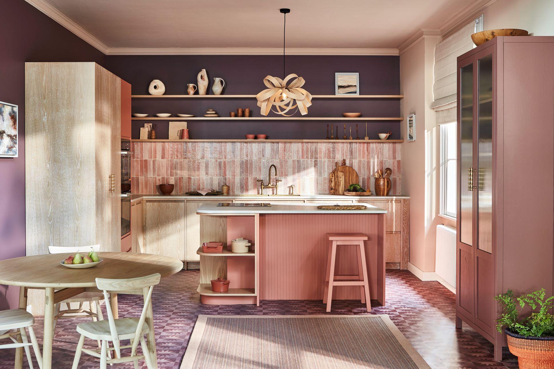 A modern kitchen with a pink and purple color scheme features a central island with bar stools, wooden cabinets, open shelves with decor, and a round dining table with chairs. A unique pendant light hangs above the island. Sunlight enters through a window.