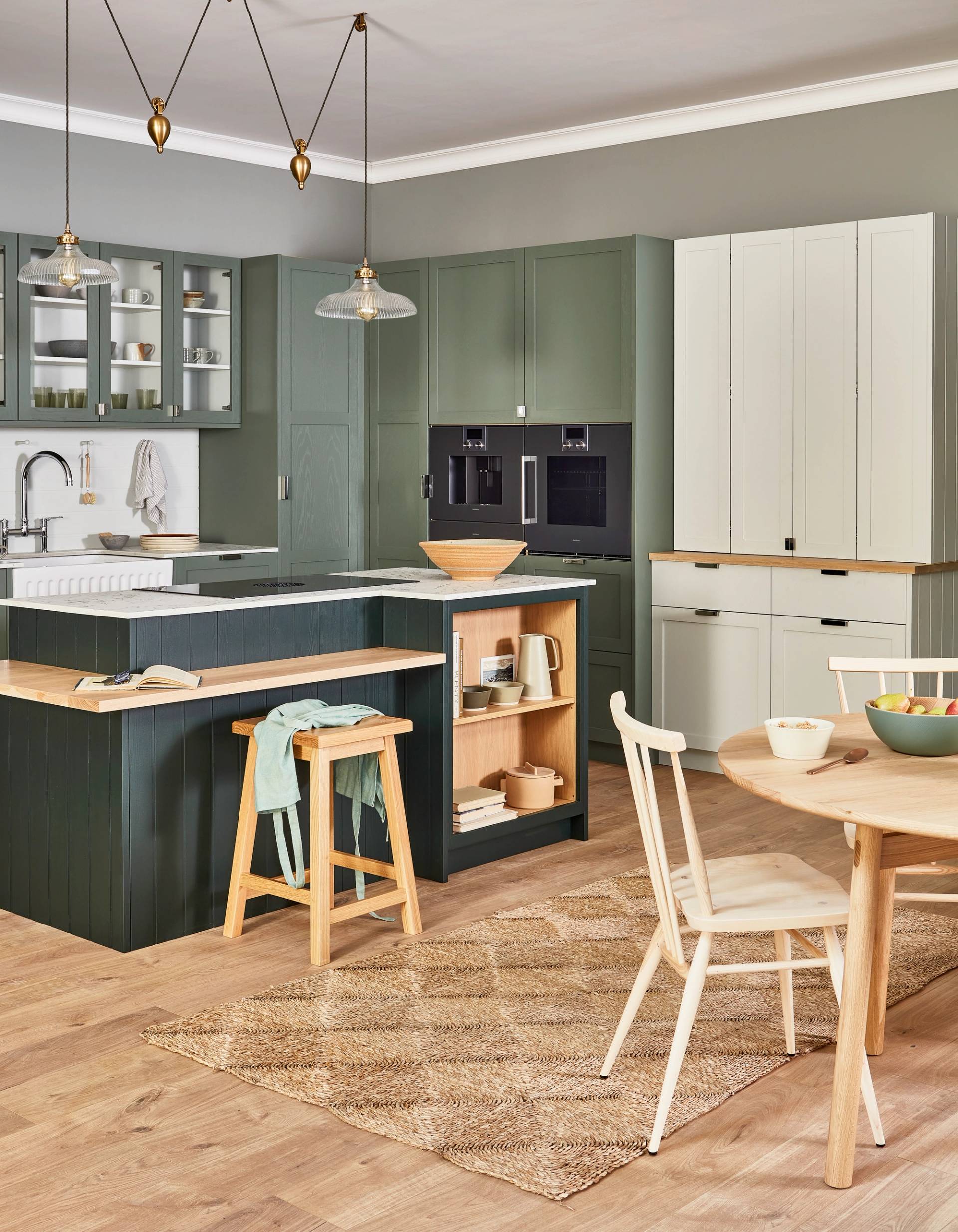 A modern kitchen with a dark green island featuring a marble countertop and wooden shelves. A tall wooden stool with a mint-green cloth draped over it stands nearby. The floor is light wood with a woven rug. Kitchenware is displayed on the shelves.