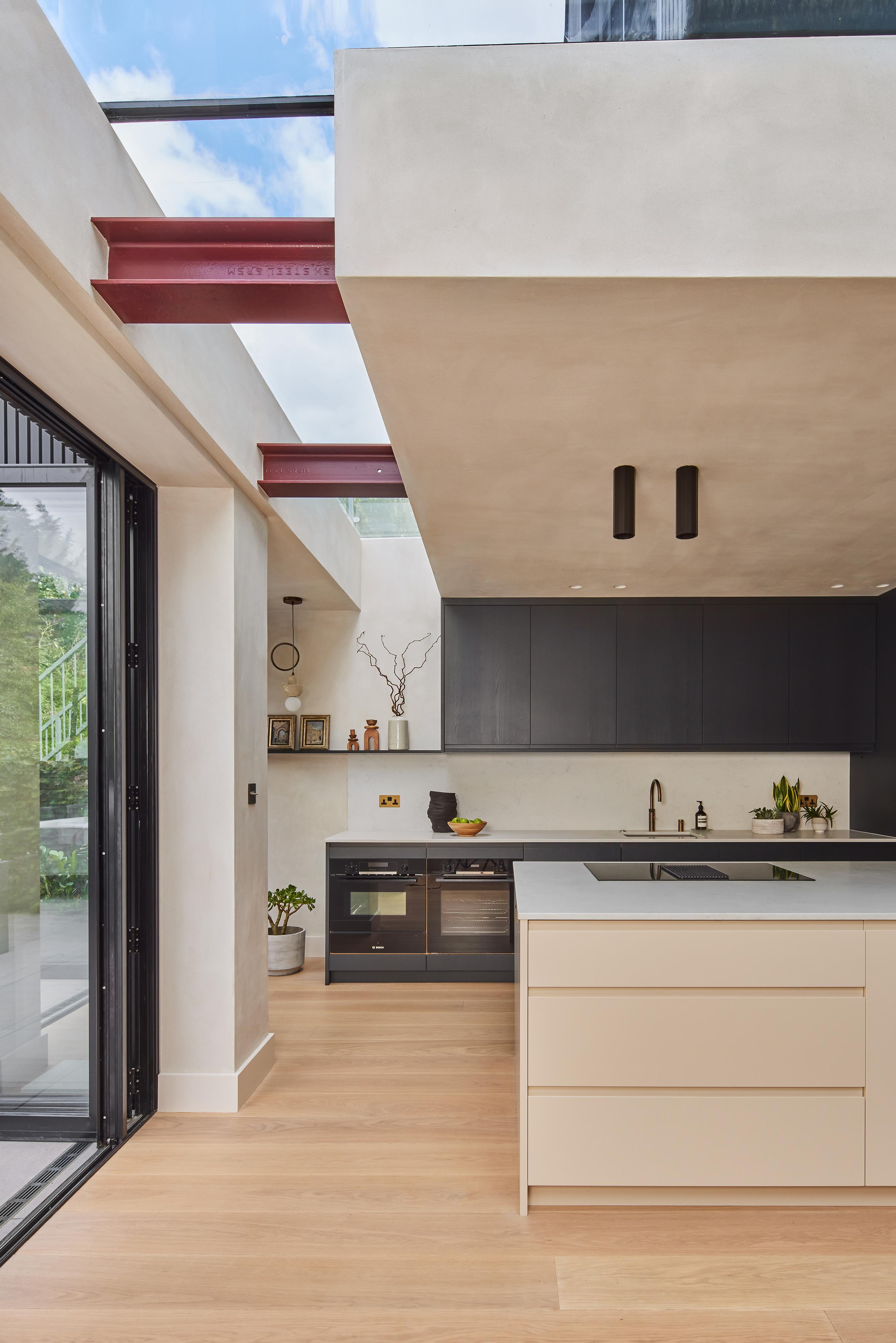 Modern kitchen with light wood flooring, a large island with beige drawers, black cabinetry, and an open shelving unit. Large glass doors open to a garden, and exposed beams are visible near the ceiling.