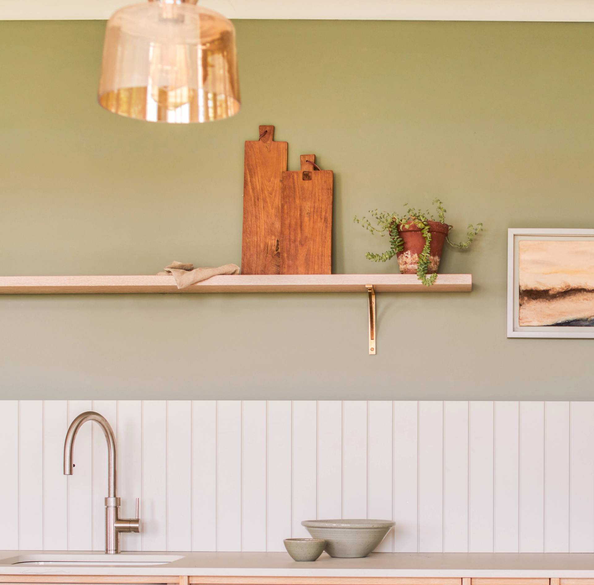 A kitchen with a minimalist design features a light green wall and a white paneling backdrop. A wooden shelf holds two cutting boards and a small plant. A modern faucet is above the sink, and two bowls sit on the counter. A framed picture is on the wall.