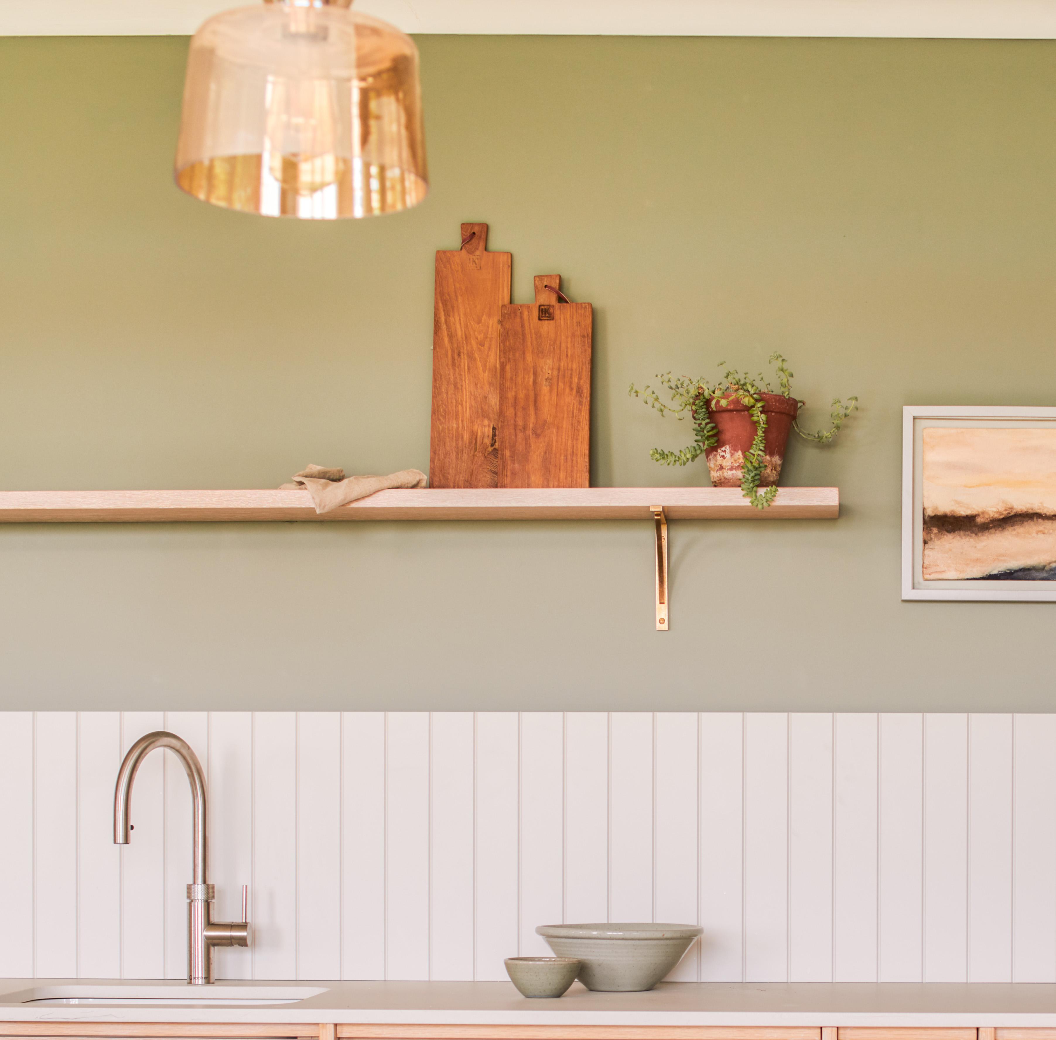 A kitchen with a minimalist design features a light green wall and a white paneling backdrop. A wooden shelf holds two cutting boards and a small plant. A modern faucet is above the sink, and two bowls sit on the counter. A framed picture is on the wall.