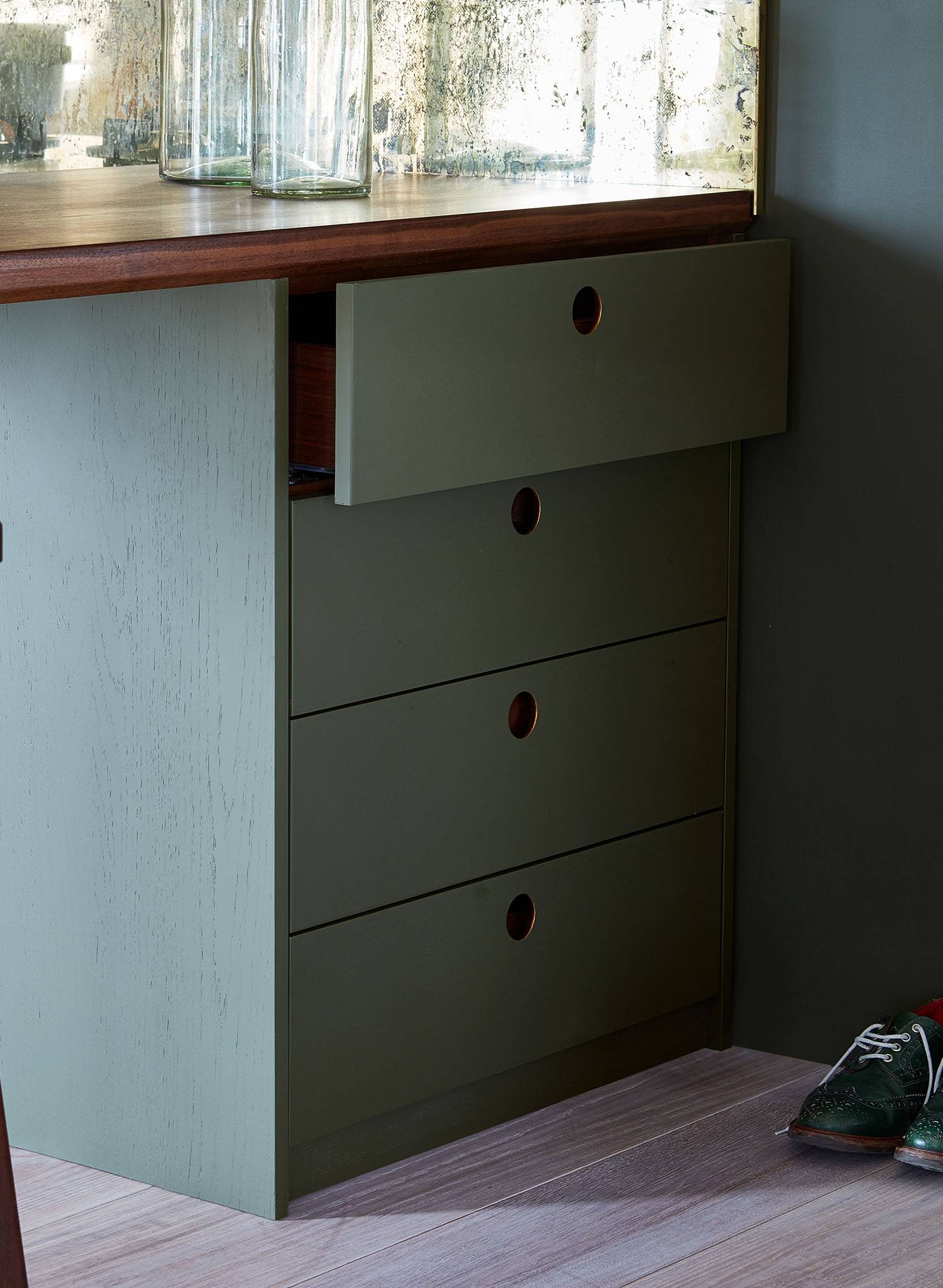 Olive green cabinet with four drawers, one partially open. Each drawer has a round cutout handle. The cabinet sits on light wood flooring, and a pair of green shoes is visible on the right side. Glass vases are on the countertop.