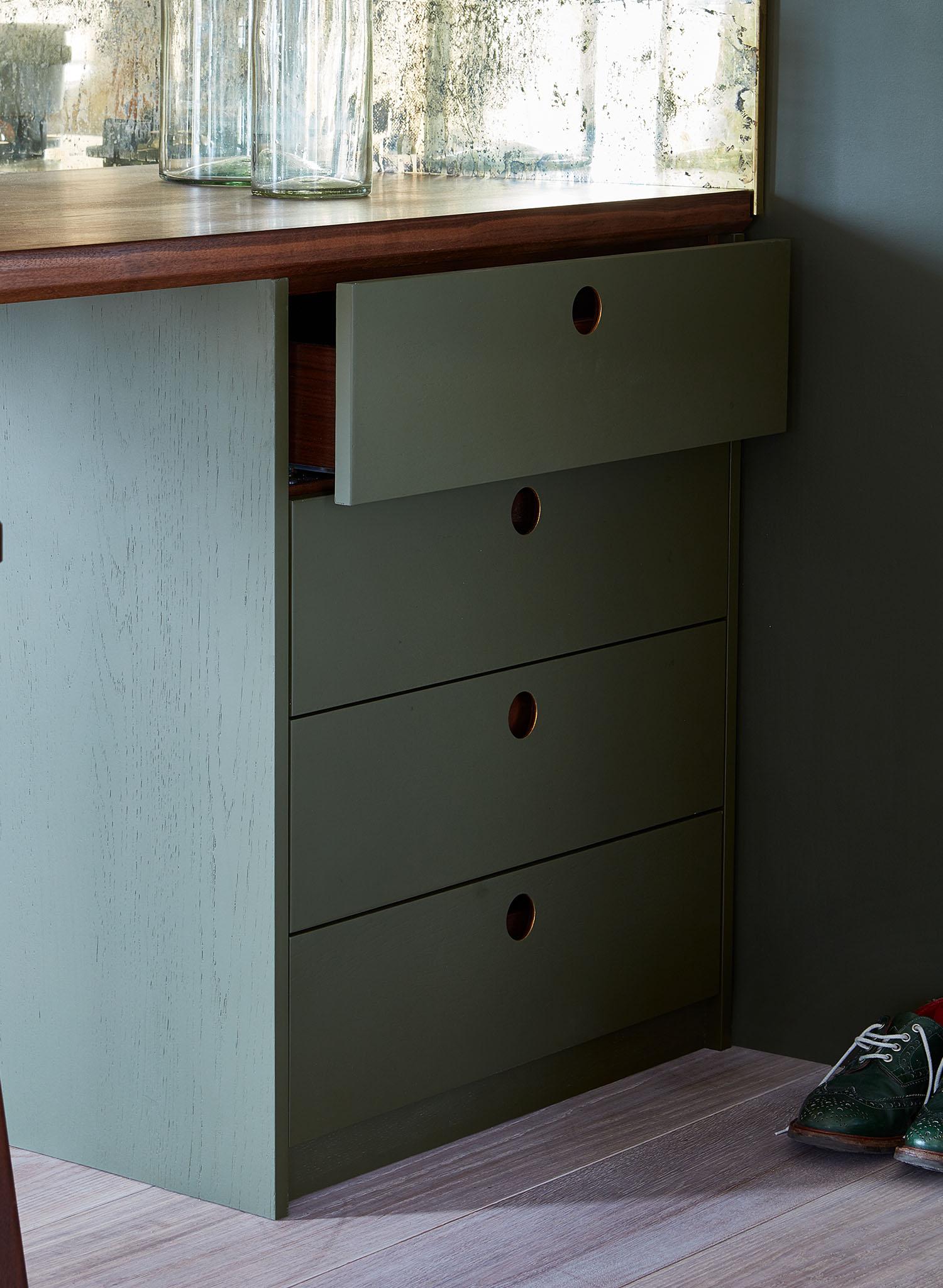 Olive green cabinet with four drawers, one partially open. Each drawer has a round cutout handle. The cabinet sits on light wood flooring, and a pair of green shoes is visible on the right side. Glass vases are on the countertop.