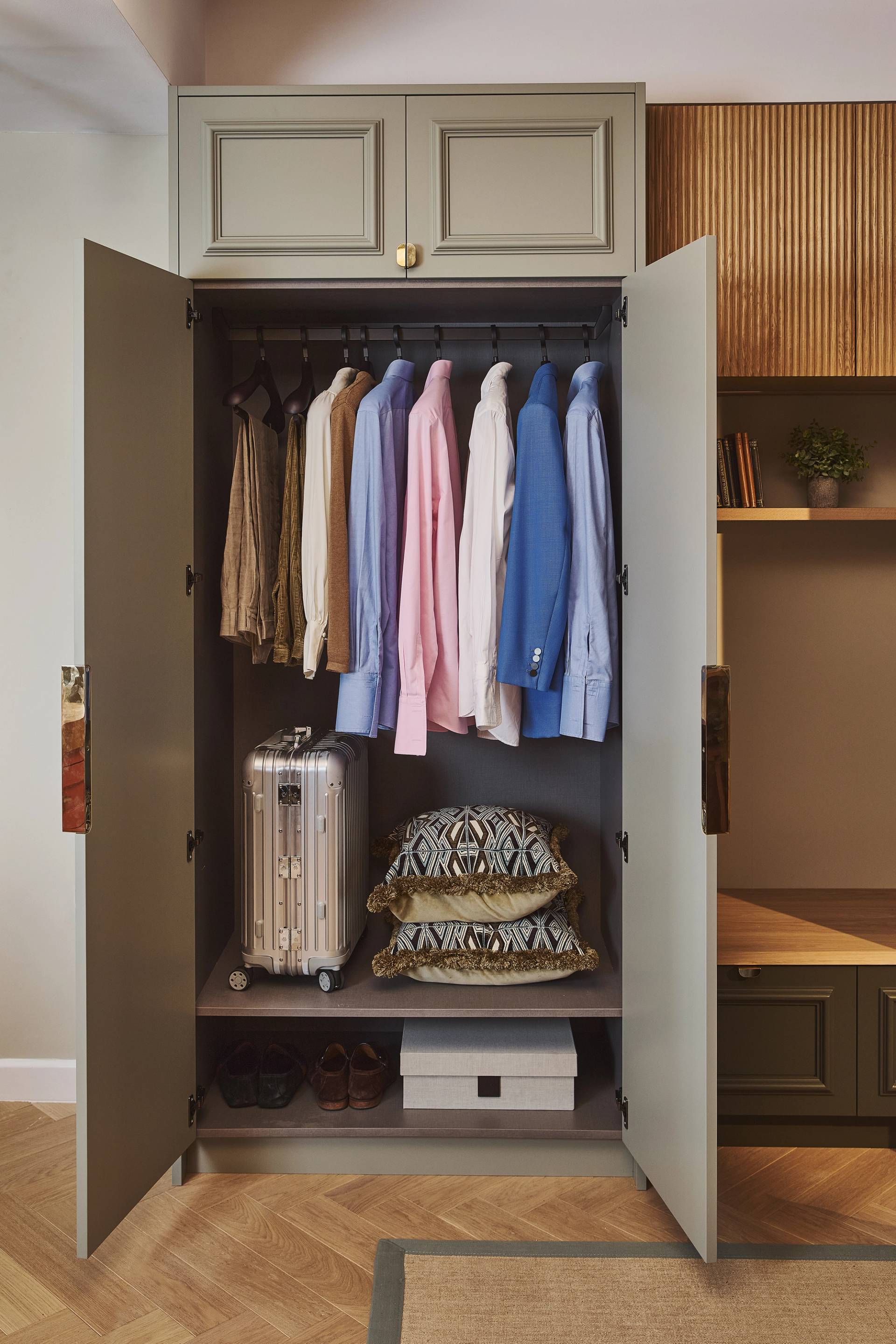 A wardrobe with open doors shows hanging shirts and jackets, a silver suitcase, patterned pillows, a few pairs of shoes, and storage boxes organized on shelves inside.