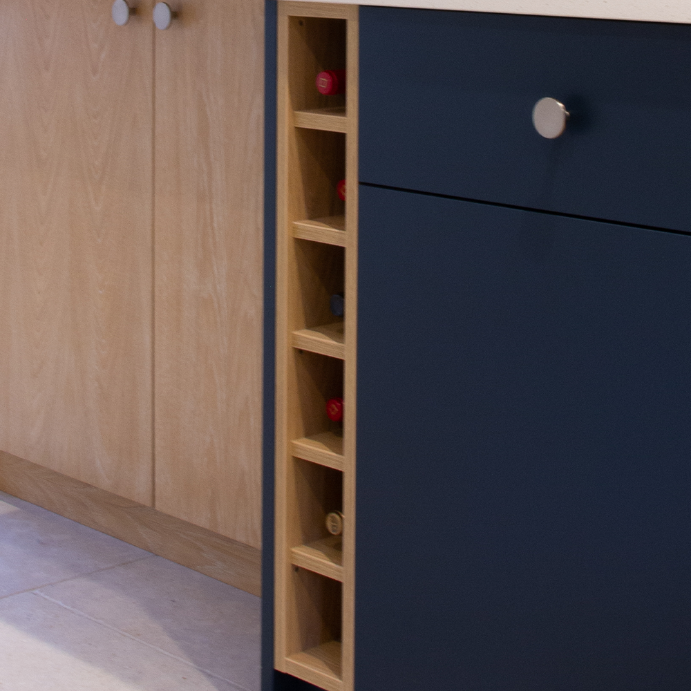 A close-up of a modern kitchen cabinet with a built-in vertical wine rack holding several bottles. The cabinet has navy blue and light wood panels with round silver handles.
