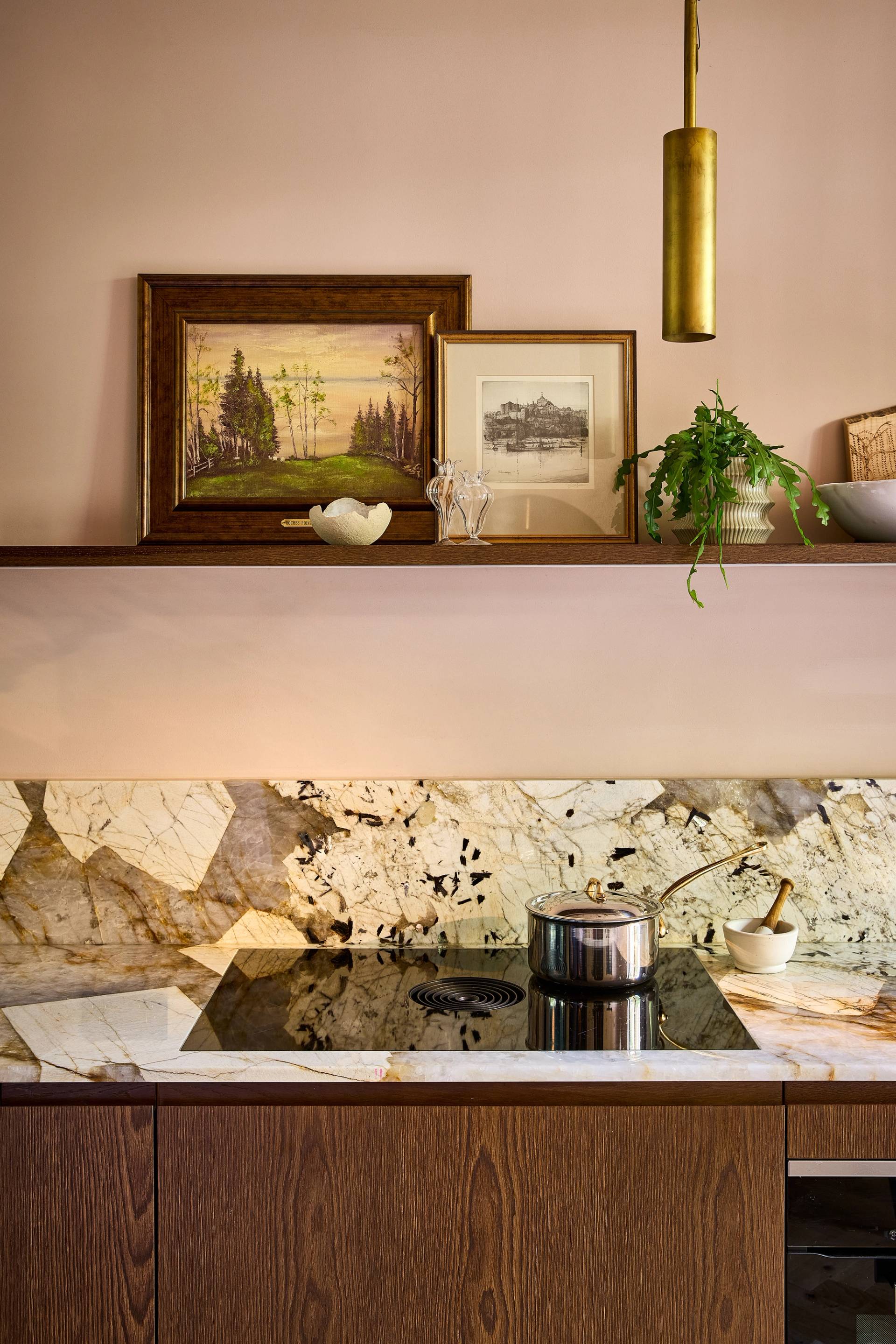 Close-up of a modern kitchen with wooden cabinets, marble backsplash, a built-in oven, cookware on the stove, open shelves holding plates, bowls, framed art, and decorative items, lit by two gold pendant lights.