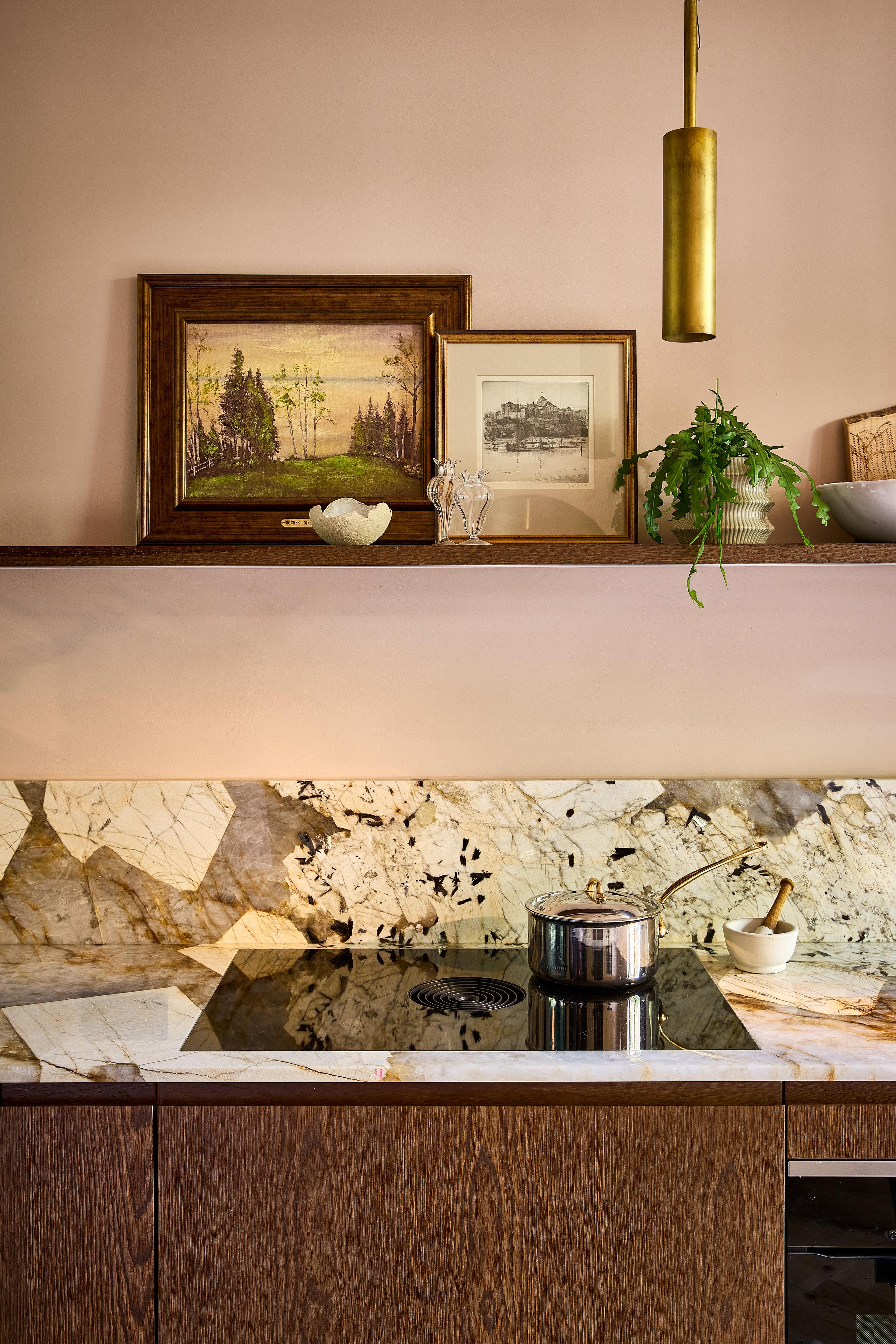 Close-up of a modern kitchen with wooden cabinets, marble backsplash, a built-in oven, cookware on the stove, open shelves holding plates, bowls, framed art, and decorative items, lit by two gold pendant lights.
