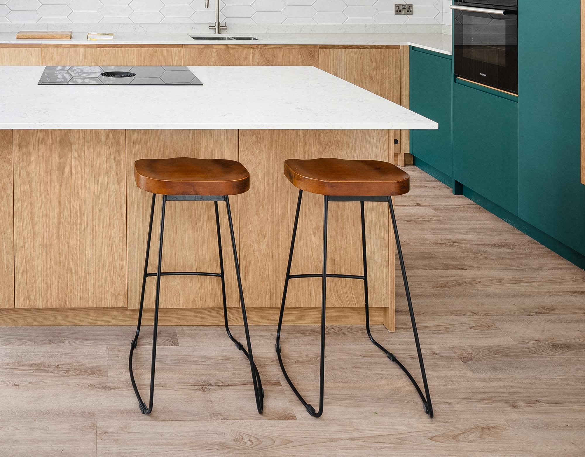 Two wooden bar stools with black metal legs are placed at a light wood kitchen island with a white countertop. The kitchen features teal cabinets and light wood flooring.
