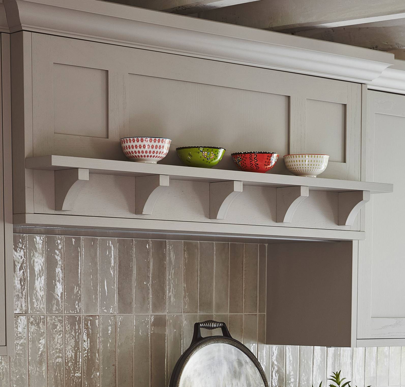A kitchen shelf holds four decorative bowls—red, green, and white—above a tiled backsplash and stove area, with a metal pan resting below. The shelf is supported by curved brackets.