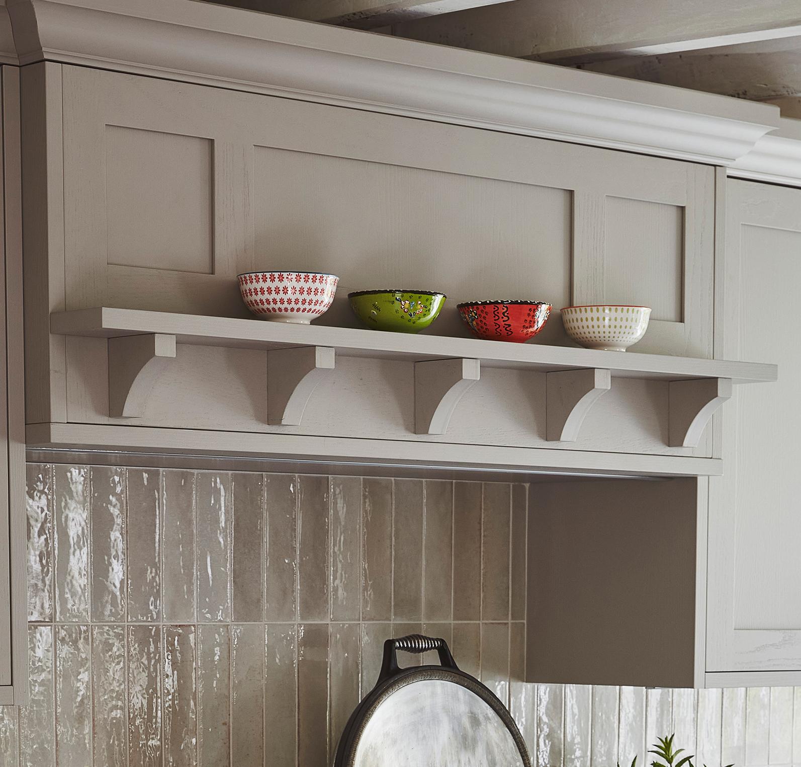 A kitchen shelf holds four decorative bowls—red, green, and white—above a tiled backsplash and stove area, with a metal pan resting below. The shelf is supported by curved brackets.