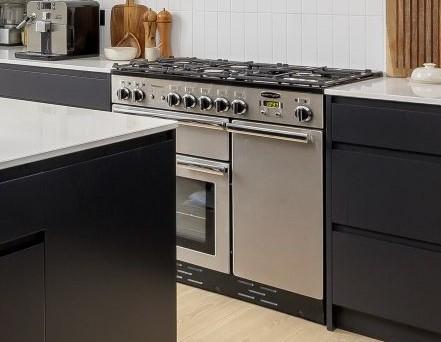 A modern stainless steel gas stove with multiple burners and two ovens is built into a sleek kitchen with black cabinets and white countertops. Cutting boards and utensils are visible on the counter.