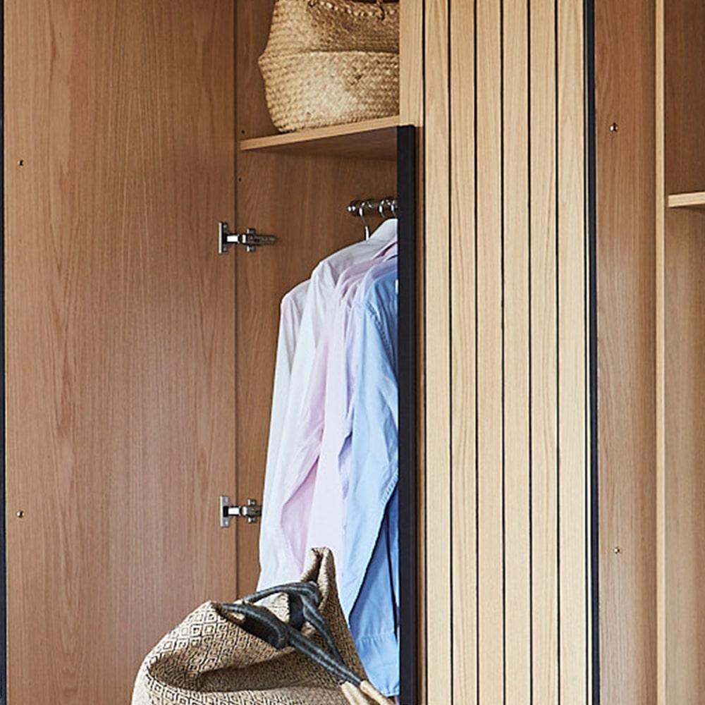 An open wooden wardrobe displaying two shirts hanging inside, with woven baskets placed on the shelves above and below.