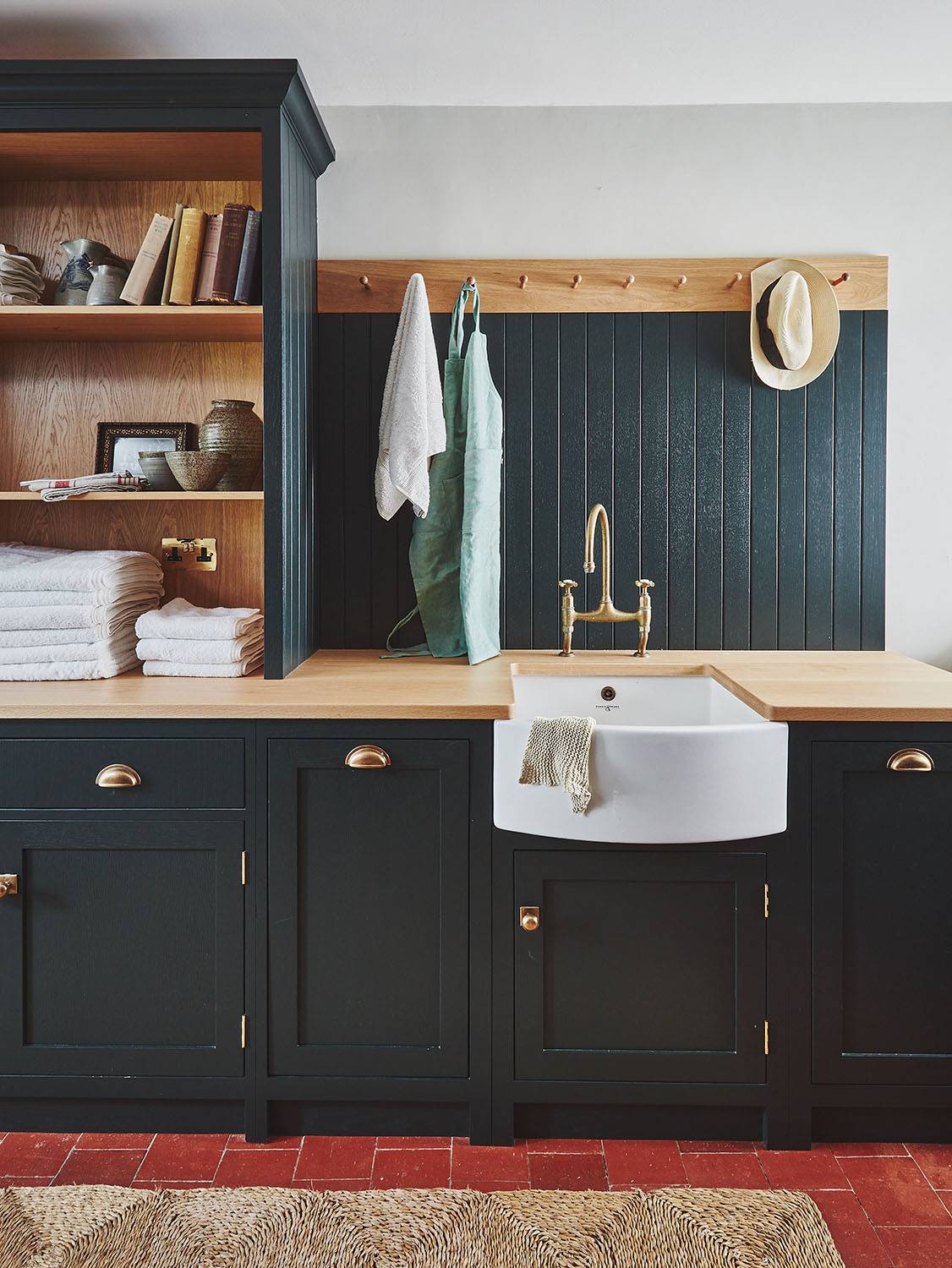 A stylish laundry room with black cabinets, a farmhouse sink, brass fixtures, folded towels, shelves with books, a hanging hat, apron, and towels, all set against a black paneled backsplash and wood countertop.