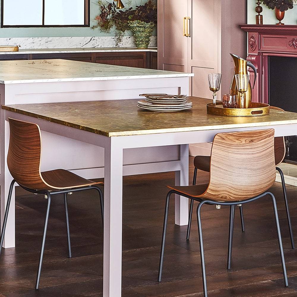A modern dining area with a wooden table, two curved wooden chairs with metal legs, and a gold tray with glassware and a pitcher. The room features dark wood floors and a red fireplace mantel in the background.