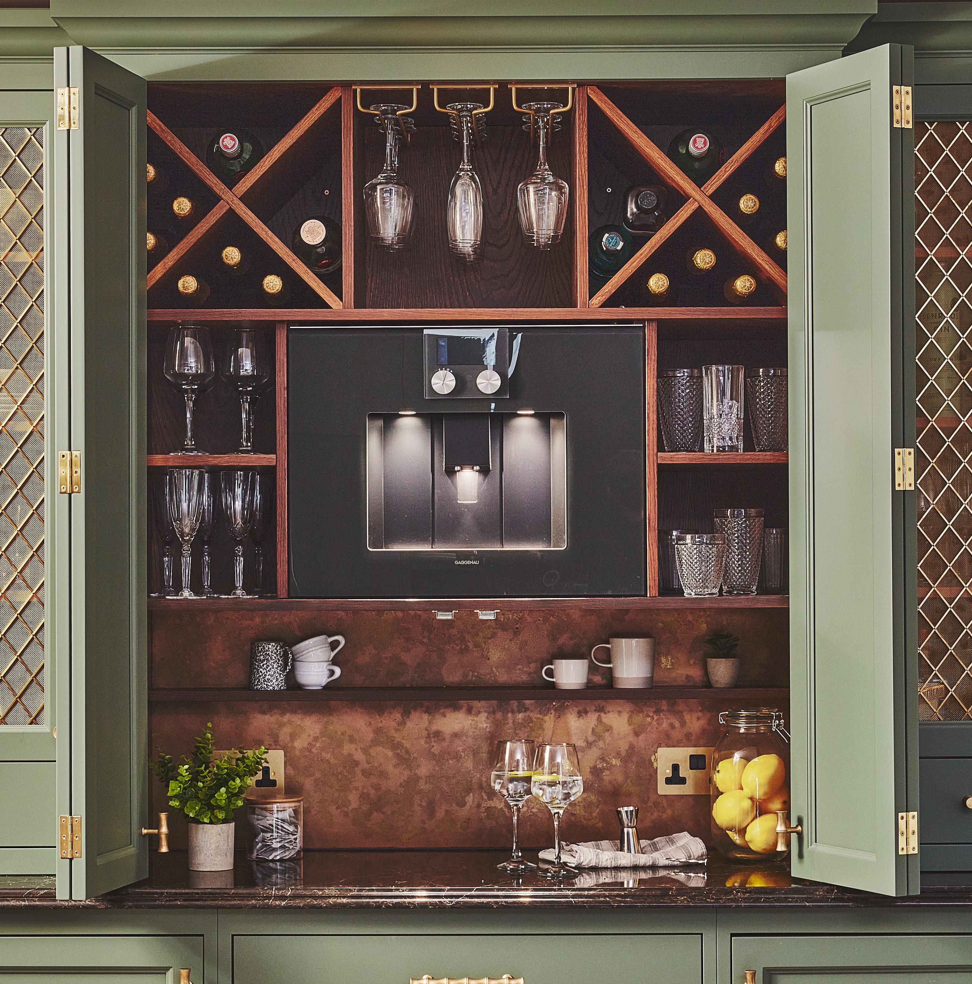 A stylish home bar with wine bottles stored in a wooden rack, glassware neatly arranged on shelves, a built-in coffee machine, and cabinets with green doors. Lemons and drinks with glasses sit on the countertop.